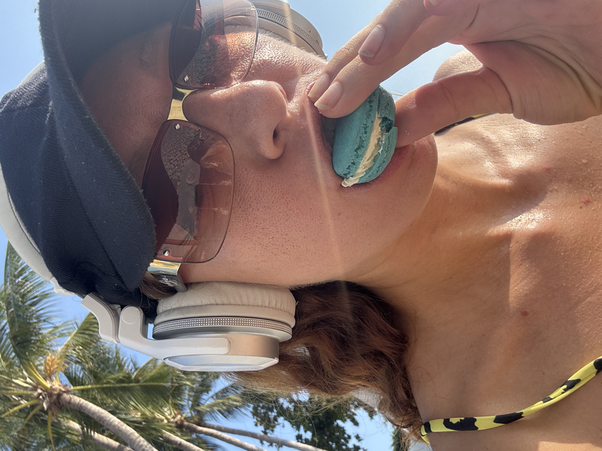 A close-up, tilted shot features a person with fair skin and brown hair, wearing sunglasses and a black and white baseball cap along with white headphones, holding a blue macaron up to their face. The person is wearing a yellow and black printed bikini top. The background reveals palm trees and a blue sky, suggesting a tropical location. Based on the prompt, the location is Baan Chaweng, Thailand.