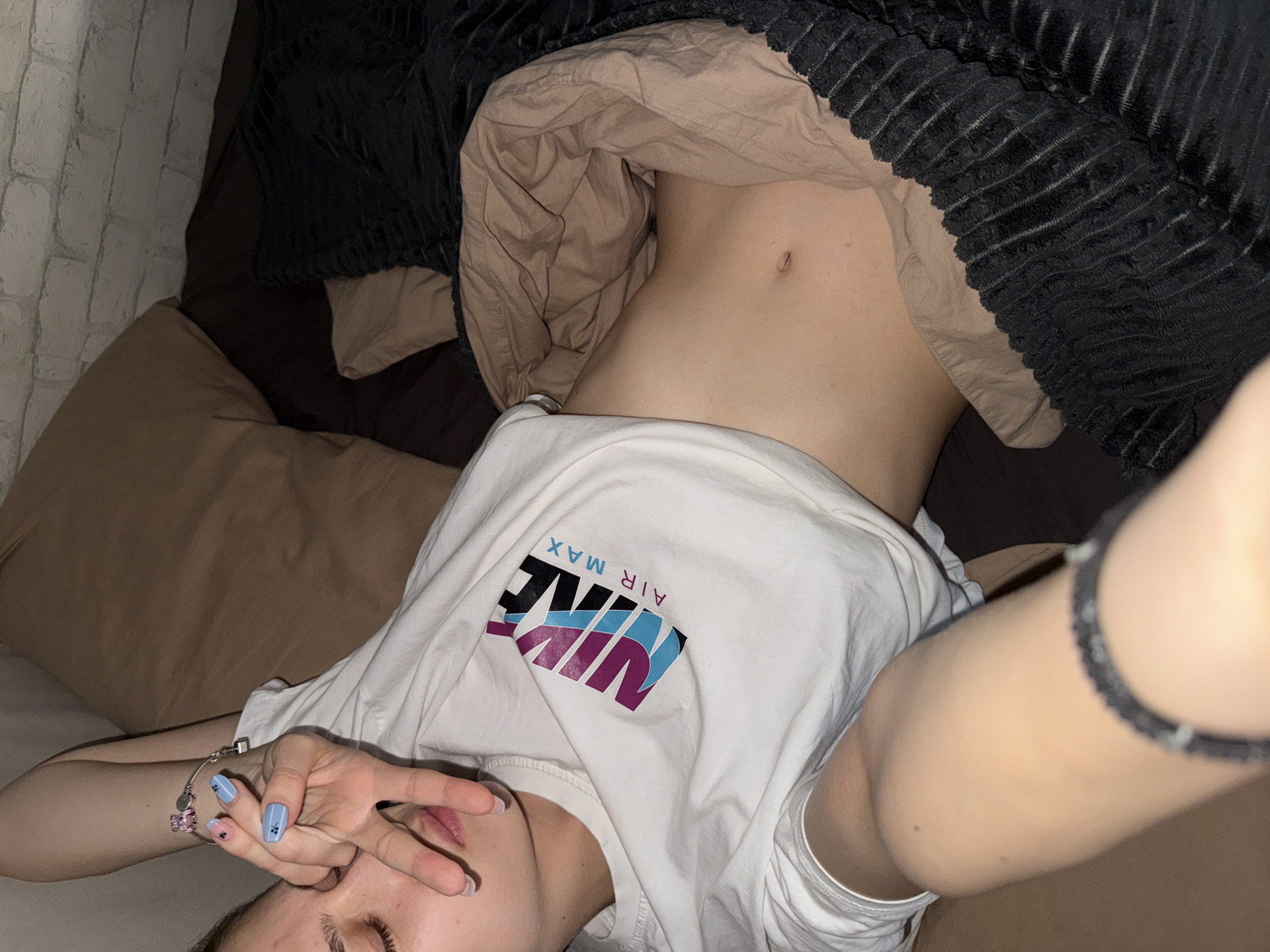 The image depicts a person lying on a bed. The individual is partially covered by a brown blanket and a black textured blanket. They are wearing a white T-shirt with a "NIKE AIR MAX" logo on the front. The person's torso is visible, revealing their stomach and navel. The individual's left arm is raised, with fingers partially obscuring their face. A bracelet adorns their left wrist, and their fingernails are painted blue. A second bracelet is seen on their right wrist. The background reveals a brown pillow and a white brick wall. The location is not discernible from the provided image.