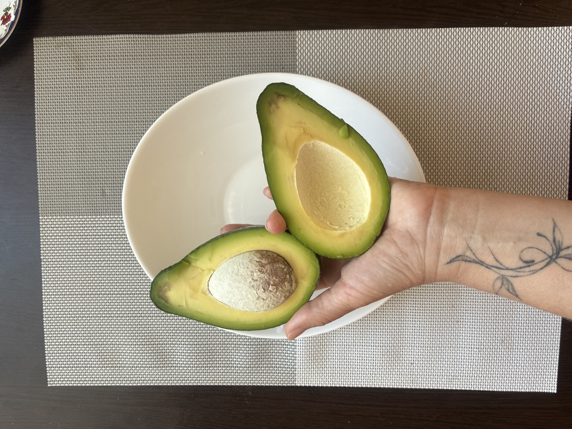 The image presents an indoor, overhead view focused on a person's hands holding two halves of a freshly cut avocado over a white bowl. The scene is set on a dark brown surface, likely a table, covered by a light gray, woven placemat.
In the center, a person's hands, with fair skin, are holding the two halves of a ripe avocado. One half, held closer to the bottom of the frame, clearly shows its large, light brown pit. The other half, held slightly above it, is empty of its pit, revealing the smooth, pale yellow-green flesh of the avocado. The flesh of both halves appears creamy and ready to eat, though the pitless half has a few small, dark blemishes near the top.
A distinctive black line-art tattoo, depicting swirling, organic shapes resembling vines or leaves, is visible on the person's right wrist and forearm. The nails are short and unpolished.
A plain, round white bowl is positioned underneath the avocado halves. In the top left corner of the image, a small portion of another decorative plate or bowl with a floral pattern in red and blue is partially visible and out of focus. The lighting suggests natural daylight. No visible text is present. The context provided indicates the location is Sochi, Russia.
