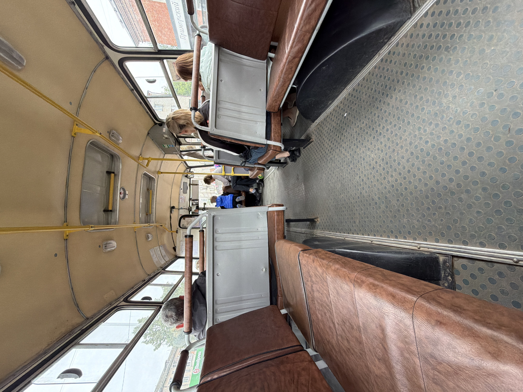 The image captures the interior of a public bus in Chaikovsky, Russia. Several passengers are seated, with their backs mostly turned towards the camera. The scene depicts the typical atmosphere of a bus journey, with people traveling from one place to another. The bus features rows of brown, leather-like seats and metal handrails for passengers to hold onto. The floor is covered with a patterned rubber matting, and the windows offer a glimpse of the outside world, suggesting it's daytime. The lighting within the bus appears to be natural, coming from the windows. There is some visible text on a sign outside the window, possibly indicating a bus stop or route, and another sign on the side of the bus that seems to be an advertisement. The overall impression is of a mundane, everyday commute.