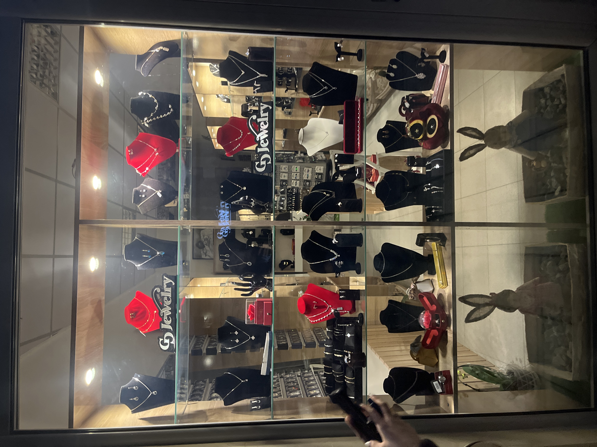 The image shows the well-lit display window of "G Jewelry" in Goce Delcev, Bulgaria. The window is divided into sections displaying various pieces of jewelry on black or red busts. There are necklaces and earrings on display. There is a stuffed bunny in the display. The reflection of someone holding a dark object, potentially a phone, is visible in the bottom right corner of the window. The store has a clean, modern interior.