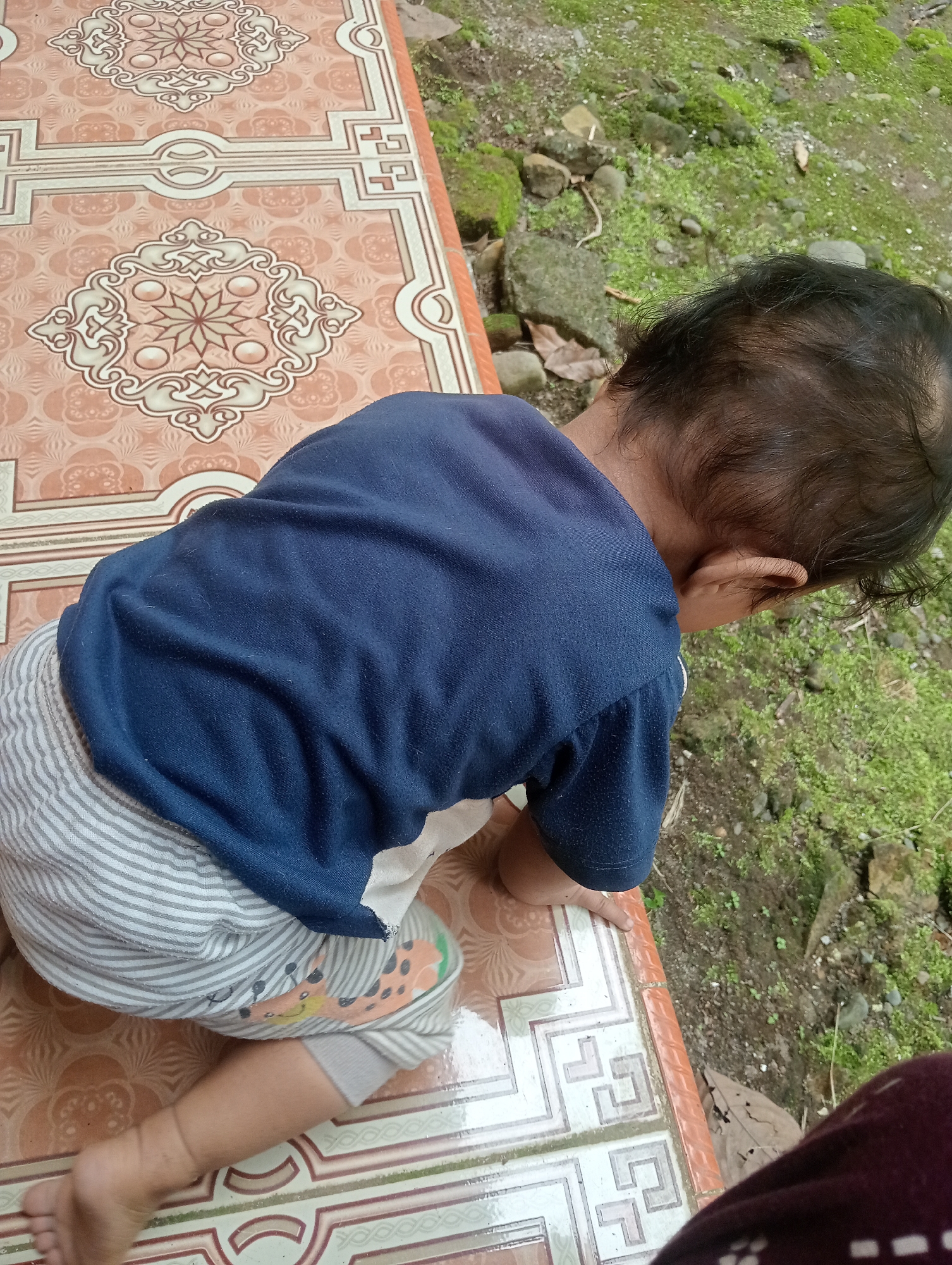 The image shows a close-up, high-angle shot of a young child from behind, kneeling on a patterned tile surface. The child is wearing a dark blue t-shirt and striped pants with a giraffe pattern on the left leg. The child's legs and feet are visible, with the left foot resting on the tile. The patterned tiles suggest an outdoor or semi-outdoor setting, possibly a patio or veranda, leading to a natural, slightly overgrown area with moss-covered rocks and dirt. The lighting suggests it could be daytime, possibly overcast or shaded. The overall mood is casual and domestic, capturing a moment of a child exploring their surroundings. There is no visible text in the image. The location context provided is Campago Barat, Indonesia, which aligns with the presence of natural elements and potentially local tile patterns.