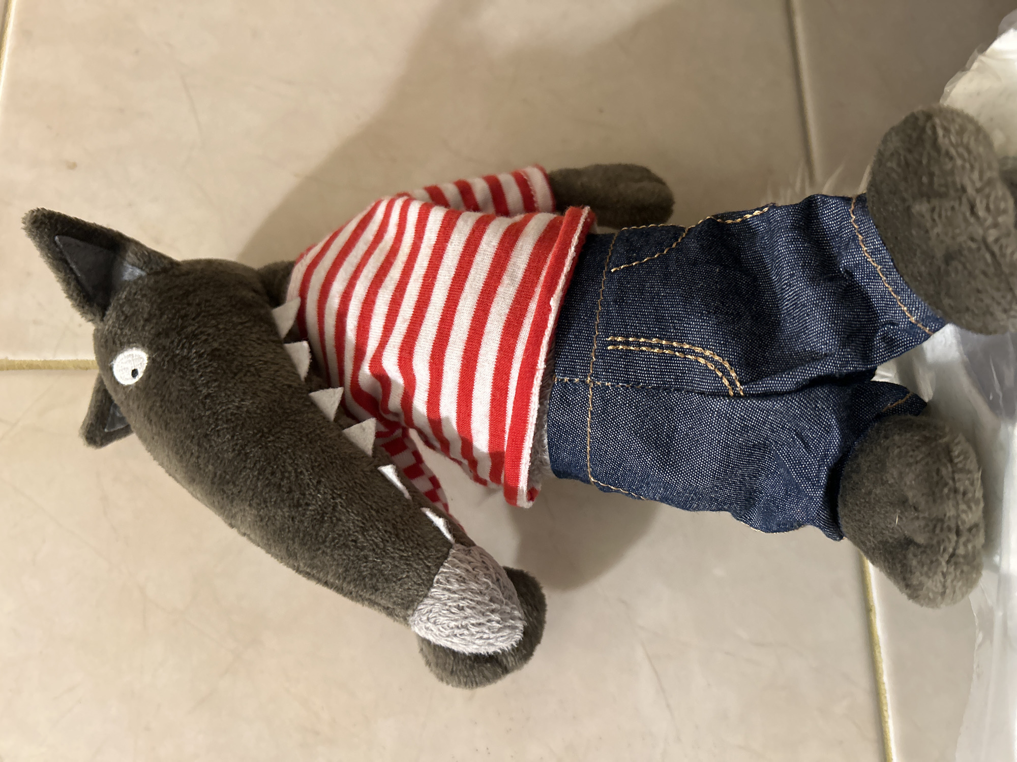 A stuffed wolf toy lies on a tiled surface. The toy is grey with a white eye and white teeth. It wears a red and white striped shirt and blue denim jeans. The environment is a light-colored tiled surface. There is no evidence to support this image being in Sofia, Bulgaria.