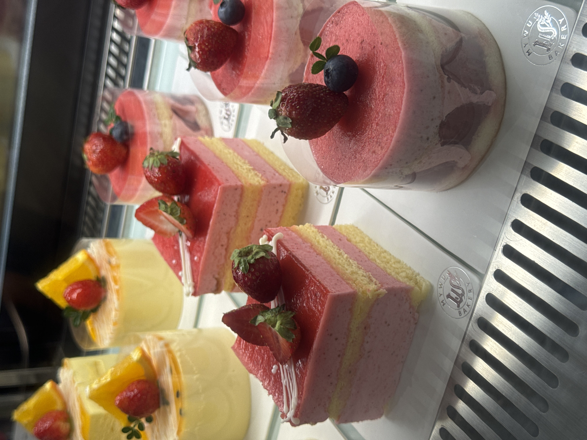 The image depicts a close-up view of pastries displayed inside a refrigerated showcase. The pastries are arranged in rows. There are cylindrical desserts, topped with fruit (strawberries and blueberries). Sliced cakes with layers of yellow cake and pink frosting, and decorated with strawberries. There are also round pastries covered in yellow icing, decorated with slices of oranges and strawberries. The pastries are arranged on glass shelves, and the shelves are located inside a refrigerated display case, with vents visible on the right side. In the upper right of the image, “My Wu Bakery” is visible. This is the My Wu Bakery, located in Phan Thiết, Vietnam. No people are visible in the photo.