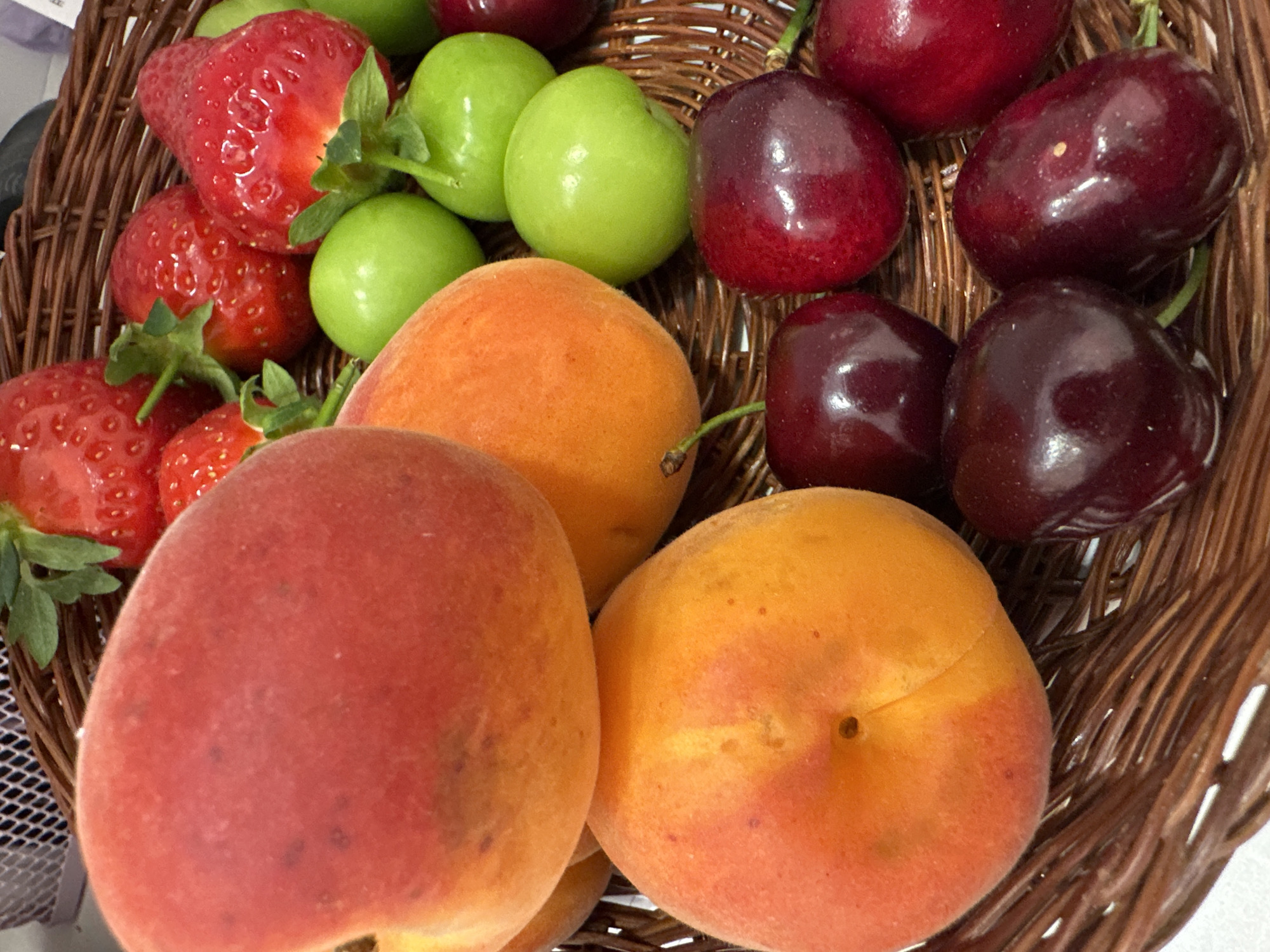 A close-up shot features a collection of fruit in a woven basket. The fruits include strawberries, green plums, apricots, and cherries. The strawberries are red with green stems, the plums are small and green, the apricots range in color from orange to reddish-orange, and the cherries are dark red. The basket is made of woven brown material. The location is unspecified but might be Sofia, Bulgaria.