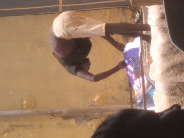 The image portrays an outdoor scene in Agege, Nigeria. A dark-skinned adult male, clad in a dark short-sleeved shirt and light-colored shorts, is bent forward at the waist. His hands are actively engaged with a rectangular object, possibly a banner or poster, featuring a prominent purple design and a printed human face, which rests on a metallic-framed structure. The background comprises a weathered, light yellow wall exhibiting significant peeling paint, with a vertical pipe affixed to its left side. A dark, vertical frame, likely a doorway, is partially visible on the far right. In the blurred foreground, the back of a dark-skinned head and shoulder of a second individual are visible in the bottom left corner. The scene is illuminated by strong natural light.