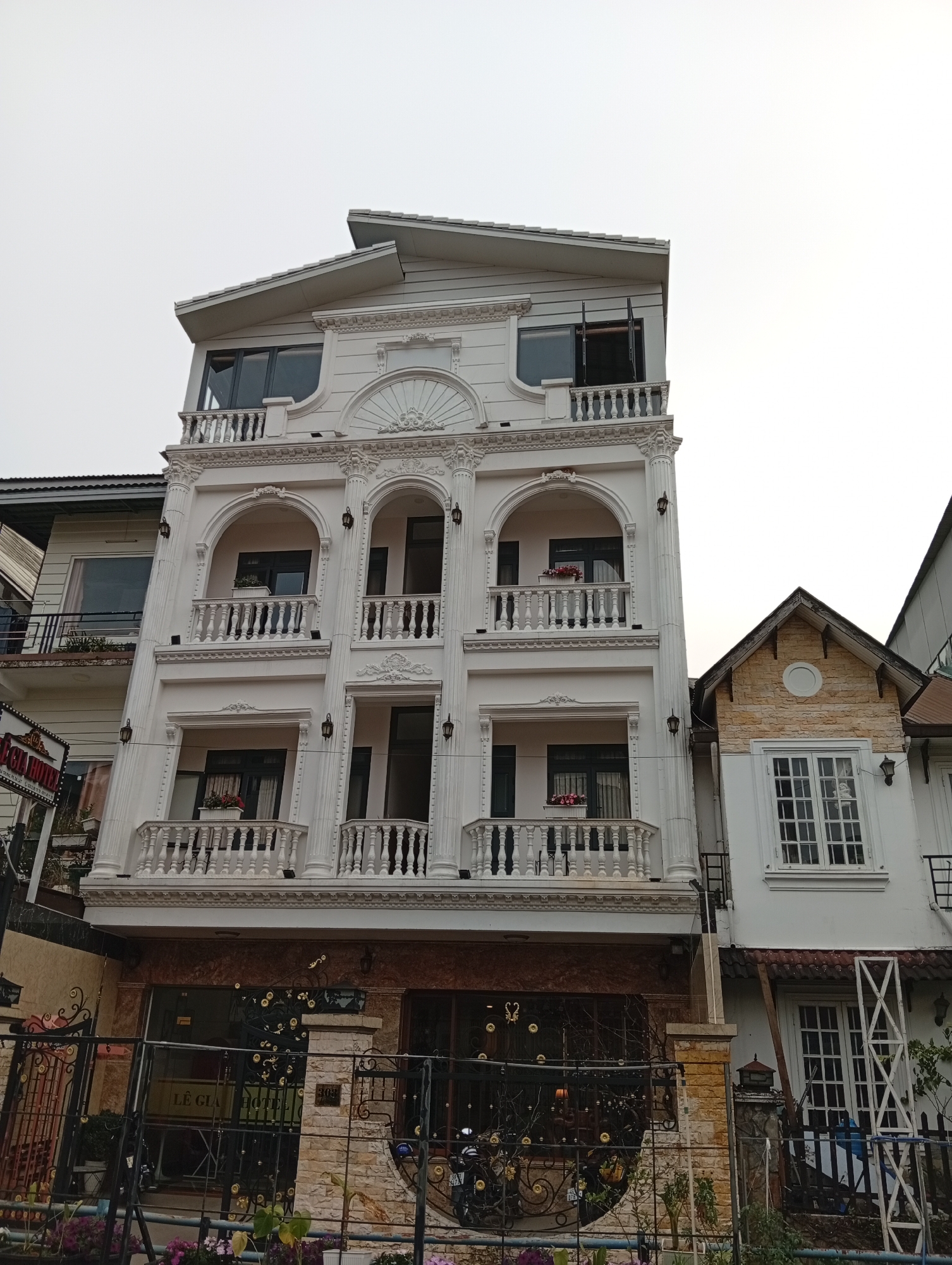 A low-angle shot depicts a four-story white building with ornate architectural details, featuring balustrades, arched windows, and decorative molding. Each story, except the ground floor, has balconies with flower boxes. The building's roof is a multi-tiered, angled design.
At street level, a wrought-iron fence encloses the property, partially obscuring the lower façade. A motorcycle is visible behind the fence. To the right, a neighboring building with a gable roof and a circular design detail is present.
The sky is overcast. The location is in the city of Xuân Hương - Đà Lạt, Vietnam.