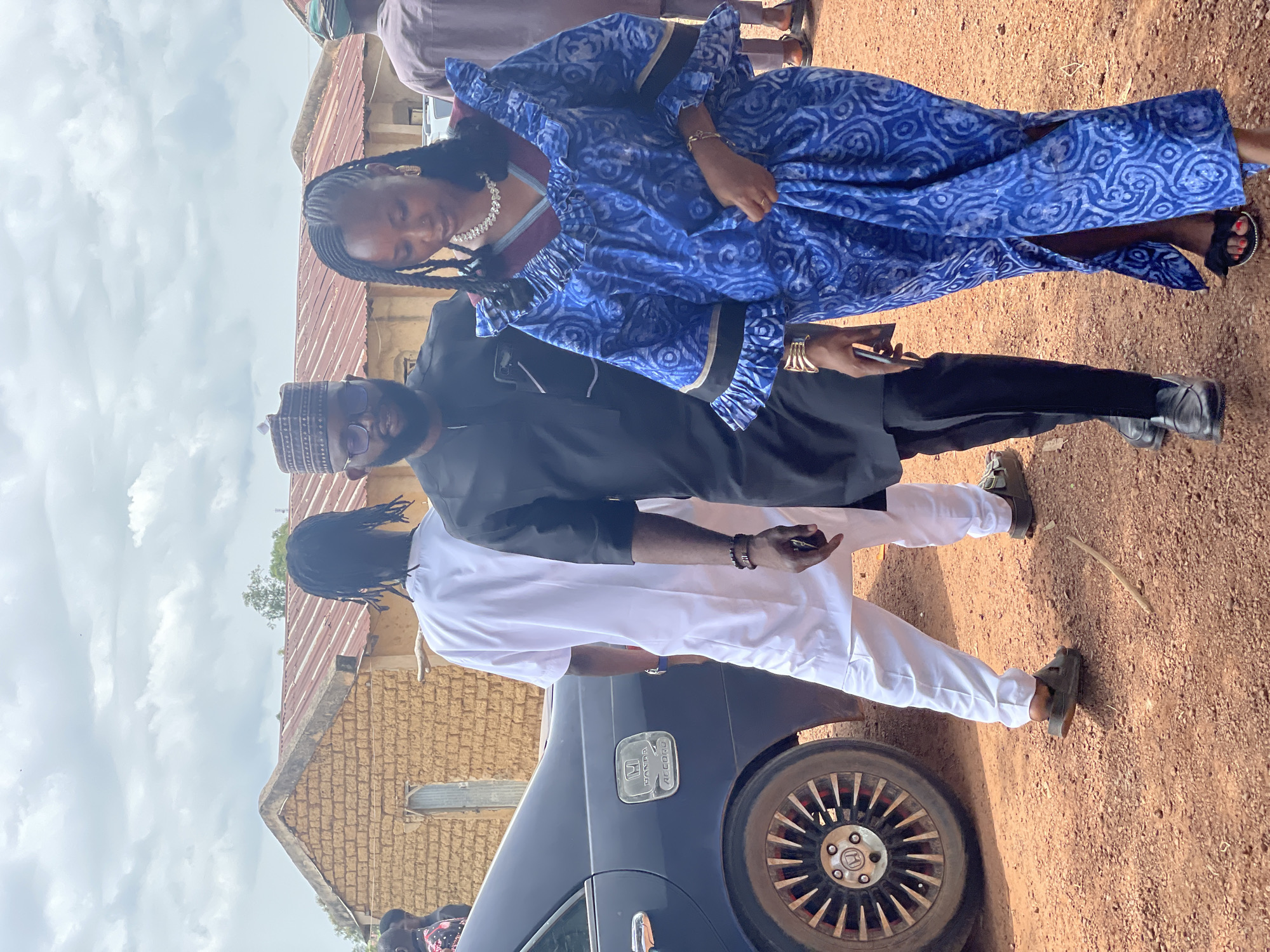 In an outdoor setting with a cloudy sky, three individuals are walking on a dirt path next to a dark blue sedan. A man in the foreground on the right wears a black traditional outfit and a cap, with eyeglasses. Next to him, a woman in a vibrant blue and white patterned dress with ruffled sleeves and a high neckline is walking, adorned with a statement necklace and bracelets. To the left of the man in black, another person dressed in a white traditional outfit is walking away from the camera. The car, identified by a "HONDA ACCORD" emblem, is parked close to the walking individuals. In the background, a single-story building with a red corrugated roof and mud-brick walls is visible. The ground is covered in reddish-brown dirt with scattered pebbles.