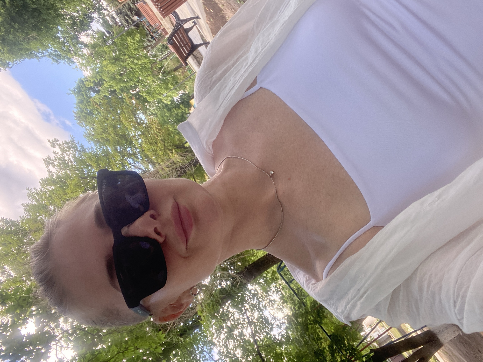 In Markivka, Ukraine, a young woman with blonde hair pulled back and a shaved undercut is captured in a selfie. She is wearing large black sunglasses, a white camisole with thin straps, and an open, lightweight white shirt. A delicate silver necklace with a small pendant rests on her collarbone. The background is a sun-dappled park with lush green trees and a bright blue sky with scattered clouds. Wooden benches are visible in the distance, suggesting a public outdoor space. The lighting indicates it is daytime, likely a sunny afternoon.