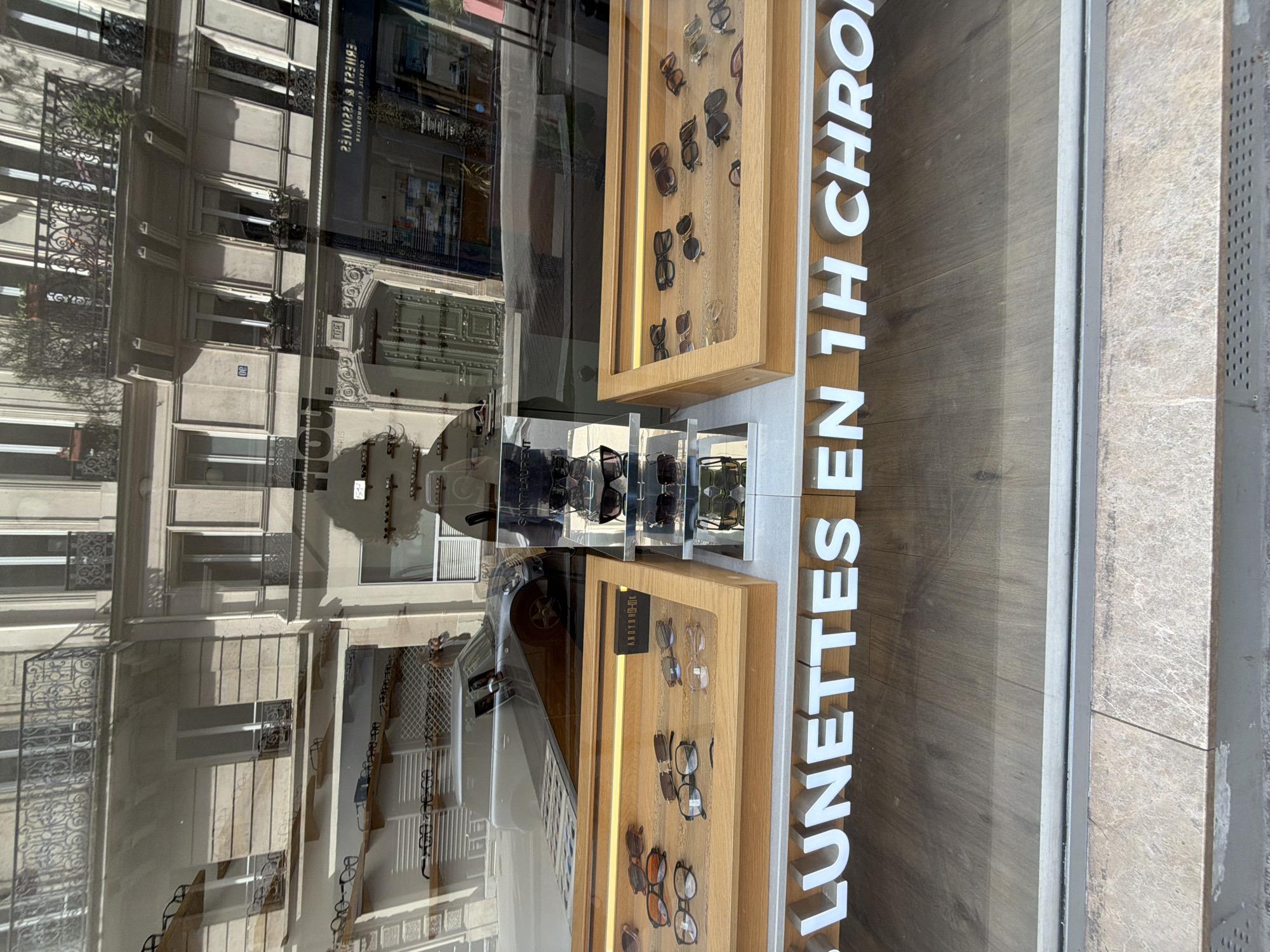 The image shows a storefront in Paris, France, with a window display of eyewear. The window has "LUNETTES EN 1H CHRONO" in large white letters. The eyewear display consists of wooden framed shelves holding sunglasses and eyeglasses. Reflected in the window are the facades of buildings across the street, typical of Parisian architecture with balconies and decorative ironwork. The buildings appear to be multi-story and well-maintained, characteristic of the urban environment. The lighting suggests a sunny day. The street is likely a commercial area, as indicated by the presence of the shop and the reflections of other businesses in the window.