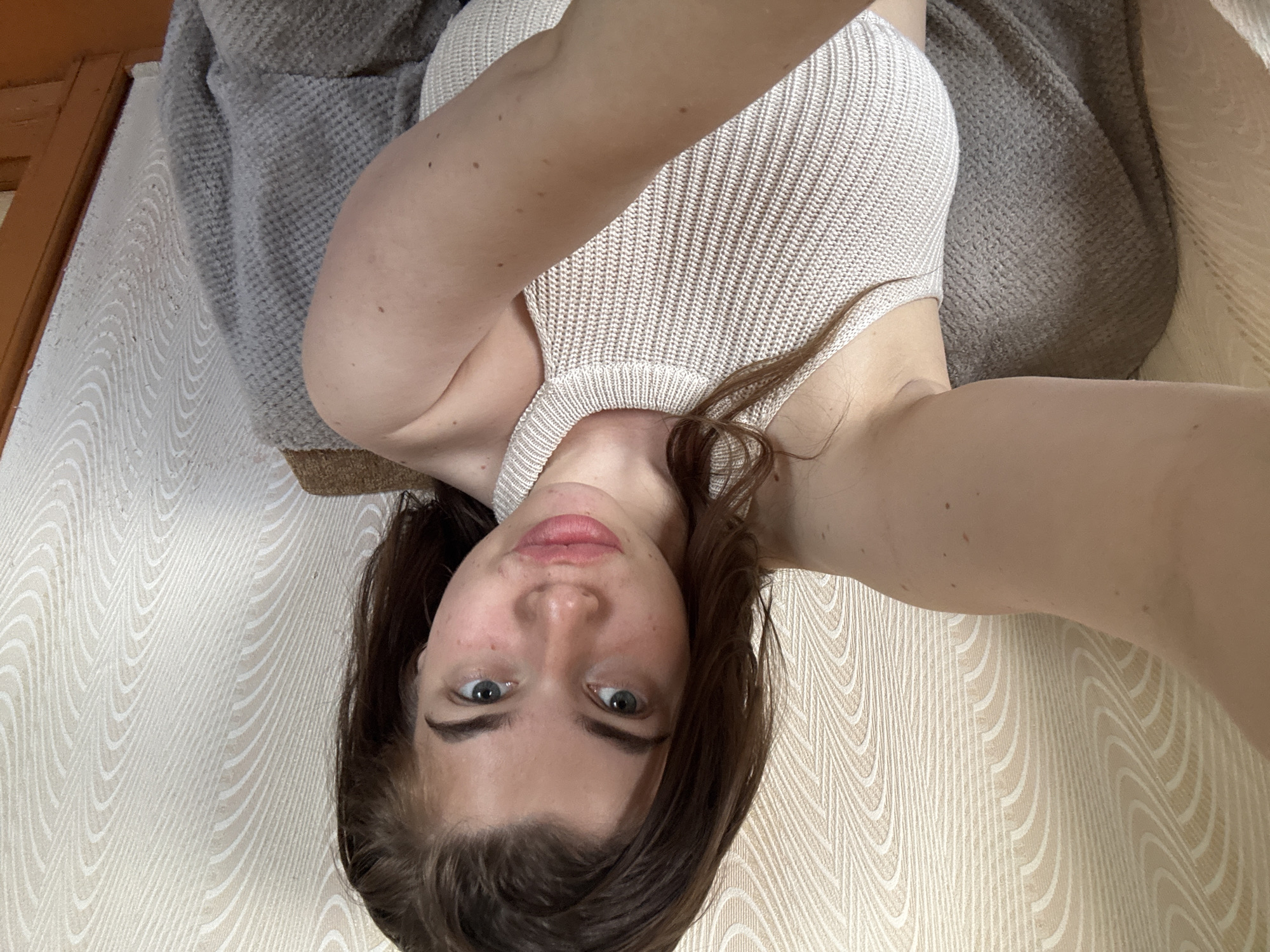 A young woman with long, brown hair and blue eyes is shown from the chest up, sitting in a chair. She is wearing a light-colored, ribbed tank top. Her left arm is extended towards the camera, with her hand out of frame. The background consists of patterned wallpaper and a hint of a doorframe. The scene appears to be indoors, likely in a residential setting. There is no indication of the woman's location in the city of Мещёры, Russia.