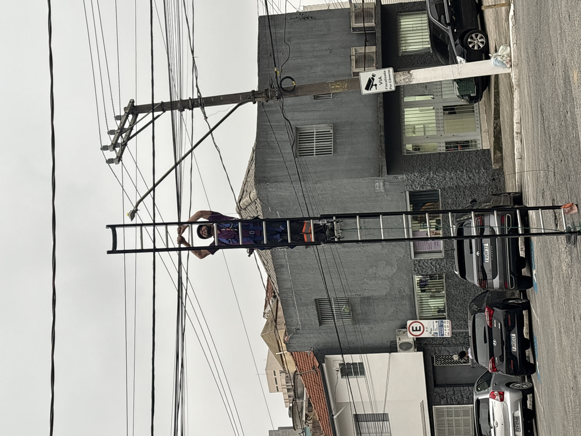 A man is standing on a tall ladder, reaching up to work with overhead wires in the city of São Paulo, Brazil. The man is wearing a dark purple shirt, a harness with blue straps, and black pants with orange reflective stripes. He has a dark cap on his head and a beard. He is gripping the sides of the ladder with both hands. The ladder is a dark color and is positioned vertically. In the background, there are several buildings, including a gray building with barred windows and a white building with a red-tiled roof. There are also power lines and a utility pole with multiple wires attached. A sign on the pole reads "VIA Fiscalizada Por Câmeras," which translates to "Area Monitored By Cameras." Cars are parked on the street in front of the buildings. The sky is overcast and gray.