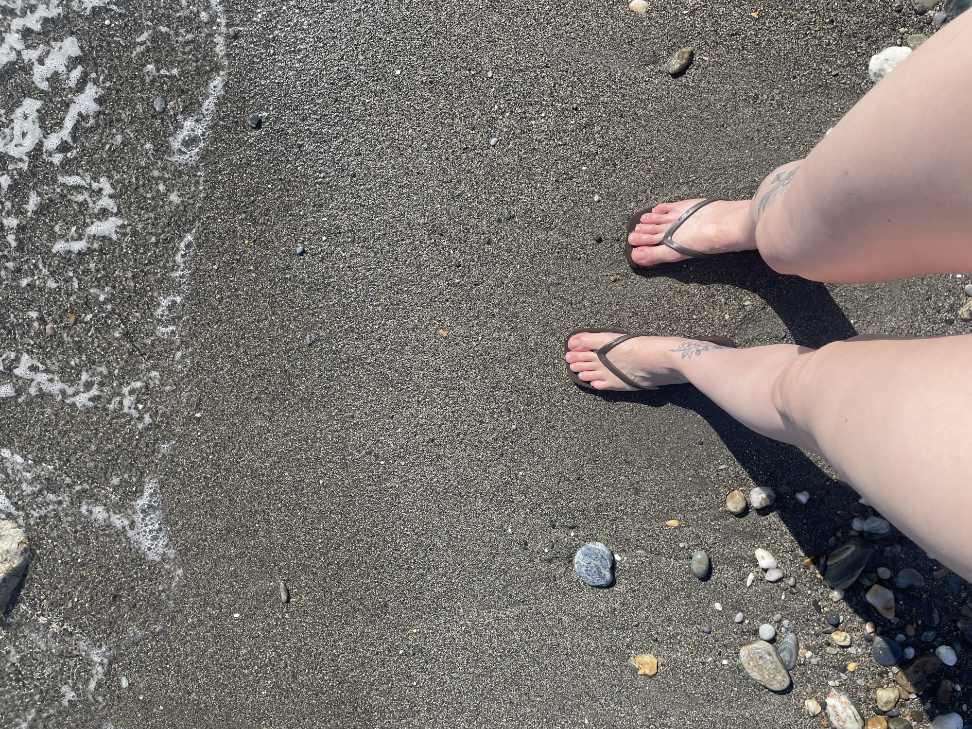 The image shows a pair of feet in dark brown flip-flops standing on a wet, dark sand beach with scattered pebbles and rocks. The water is lapping gently at the edge of the sand, creating a foamy pattern. The lighting suggests it is daytime, with a shadow cast to the left. There are subtle tattoos visible on the person's legs. The location is Unknown, Spain.