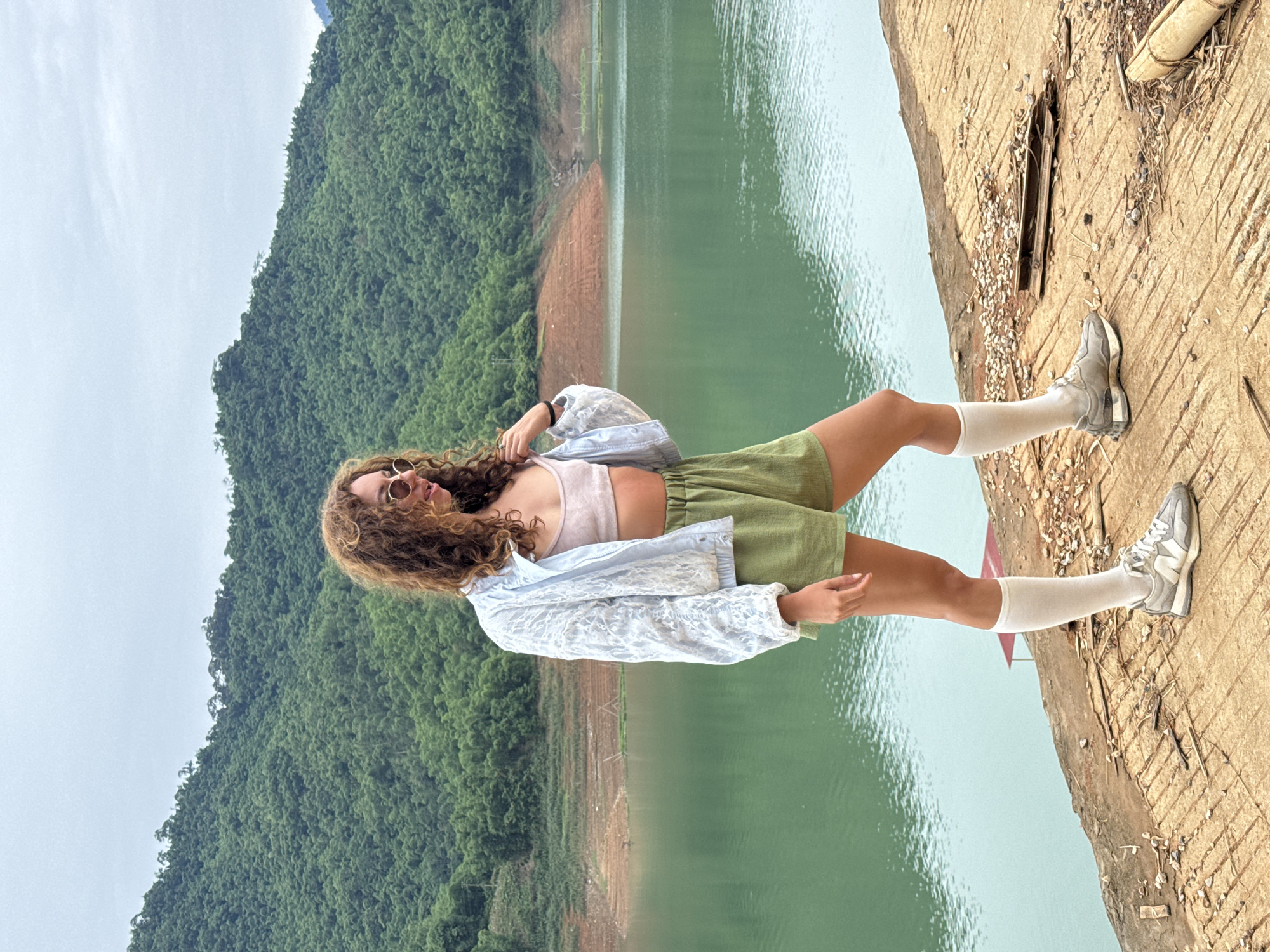 A young woman with curly brown hair and sunglasses stands on a concrete surface near a body of water with a lush green mountain range in the background. She is wearing a light pink crop top, olive green shorts, a light blue patterned jacket, white socks, and grey sneakers. The concrete surface appears weathered and is scattered with small rocks and debris. The water is a vibrant green, and the mountain slopes are densely covered with trees. The sky is overcast. This location appears to be in the vicinity of Bản Phúc, Vietnam, a region known for its natural beauty and mountainous terrain.