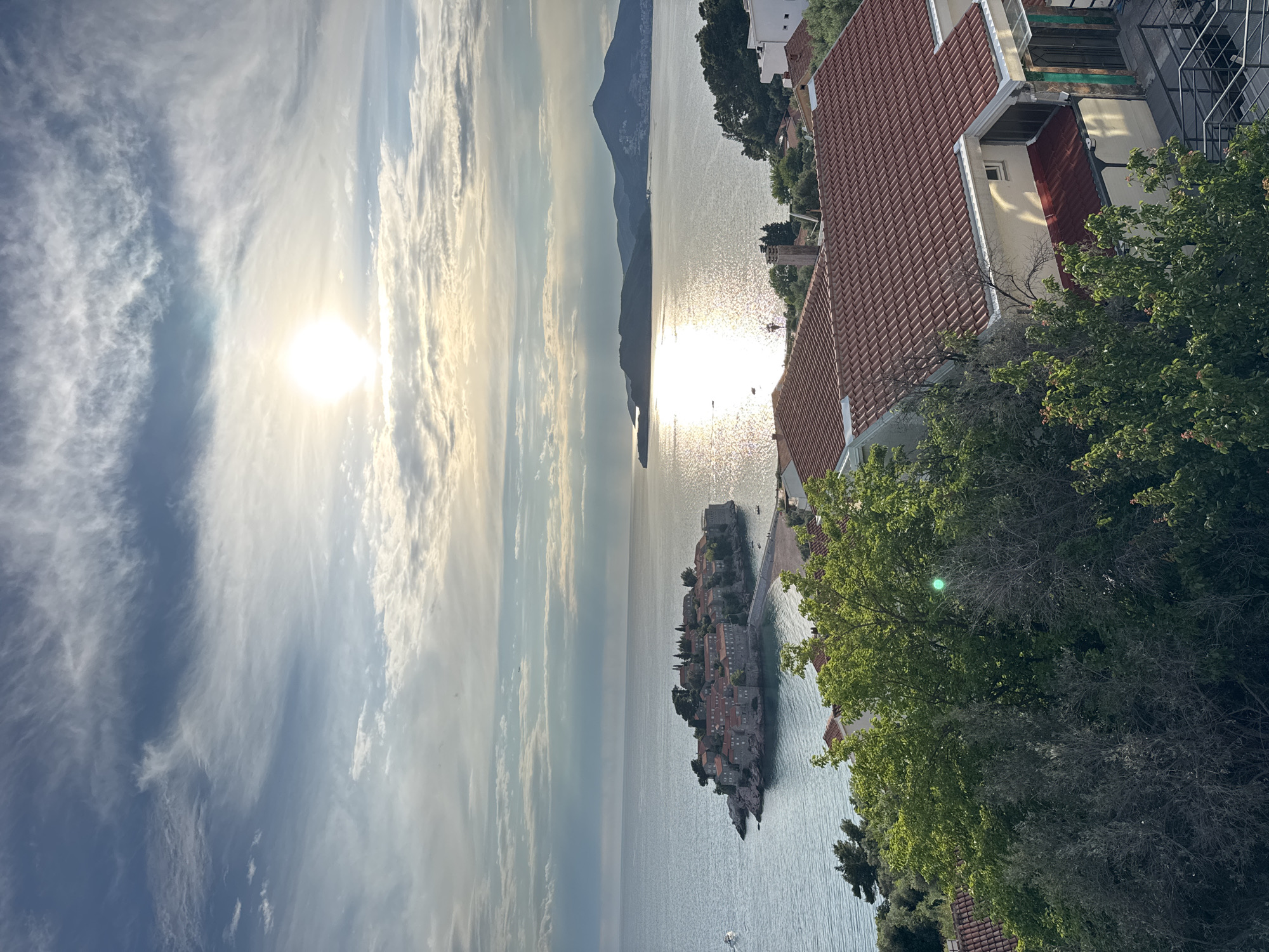 The image shows a landscape view taken from an elevated position in Sveti Stefan, Montenegro. The sun shines brightly in a partly cloudy sky above a wide expanse of calm water that reflects the sunlight. In the center, a small, densely built island topped with buildings and structures lies nestled in the water. Its terra-cotta rooftops are visible. The shoreline of Sveti Stefan is lined with trees and structures. In the foreground, the corner of a building with a red tile roof is visible, partially obscured by greenery. A mountain range can be seen in the background.