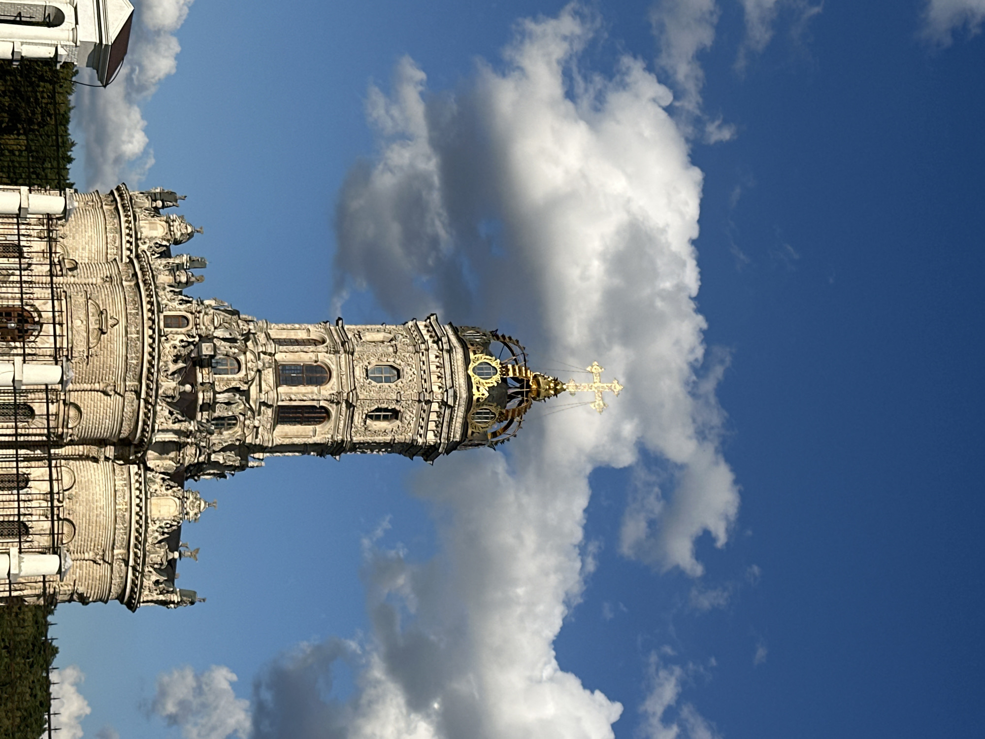 The image shows a close-up, low-angle view of the Church of the Icon of the Mother of God of All the Afflicted in Dubrovitsy, Russia. The church's intricate, baroque architecture is prominently featured, with its multi-tiered structure and ornate detailing. A golden cross crowns the top of the main tower, which is surrounded by a golden, intricate dome structure. The church is set against a vibrant blue sky with scattered white clouds. In the lower portion of the image, a black metal fence and some greenery are visible, along with a portion of a white structure to the left, possibly an entrance gate or another building. The lighting suggests it is daytime, with the sun illuminating the church and casting shadows. There are no people visible in the frame.
