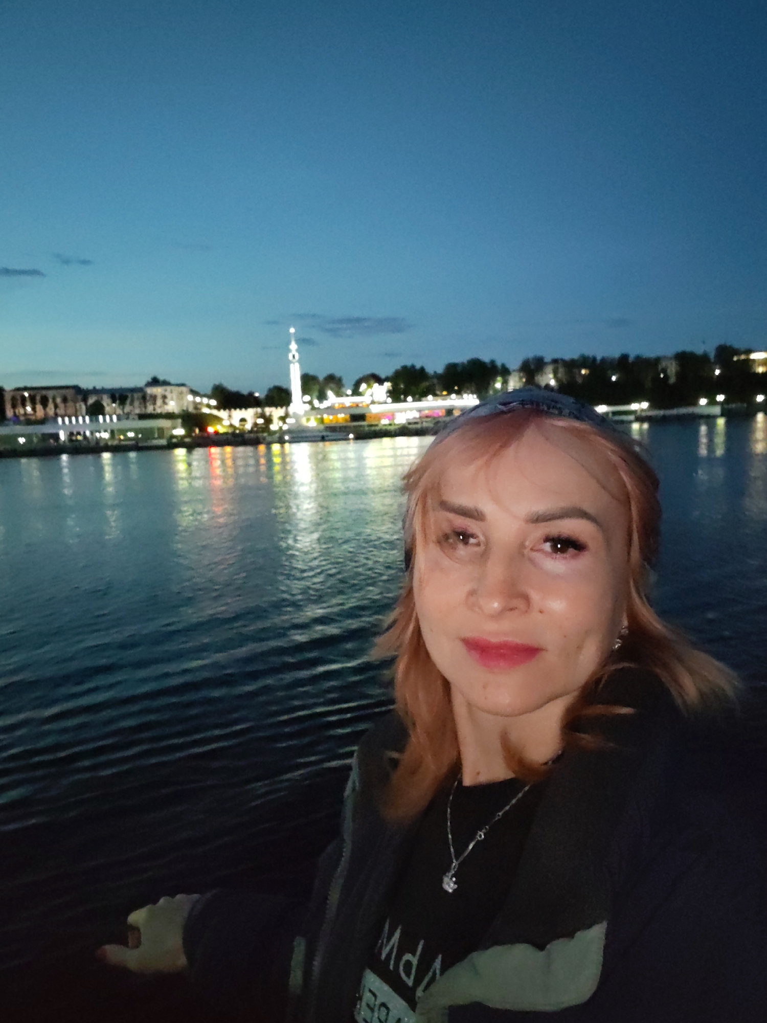 A selfie taken at dusk shows a woman with blonde hair, wearing a dark jacket over a t-shirt and a silver necklace. She is standing near a body of water, likely a river or bay, with a city skyline visible in the background. The city lights are reflected in the water. The sky is a deep blue, indicating twilight. The environment suggests an urban setting in Шебунино, Russia, with architectural structures and illuminated buildings lining the waterfront.