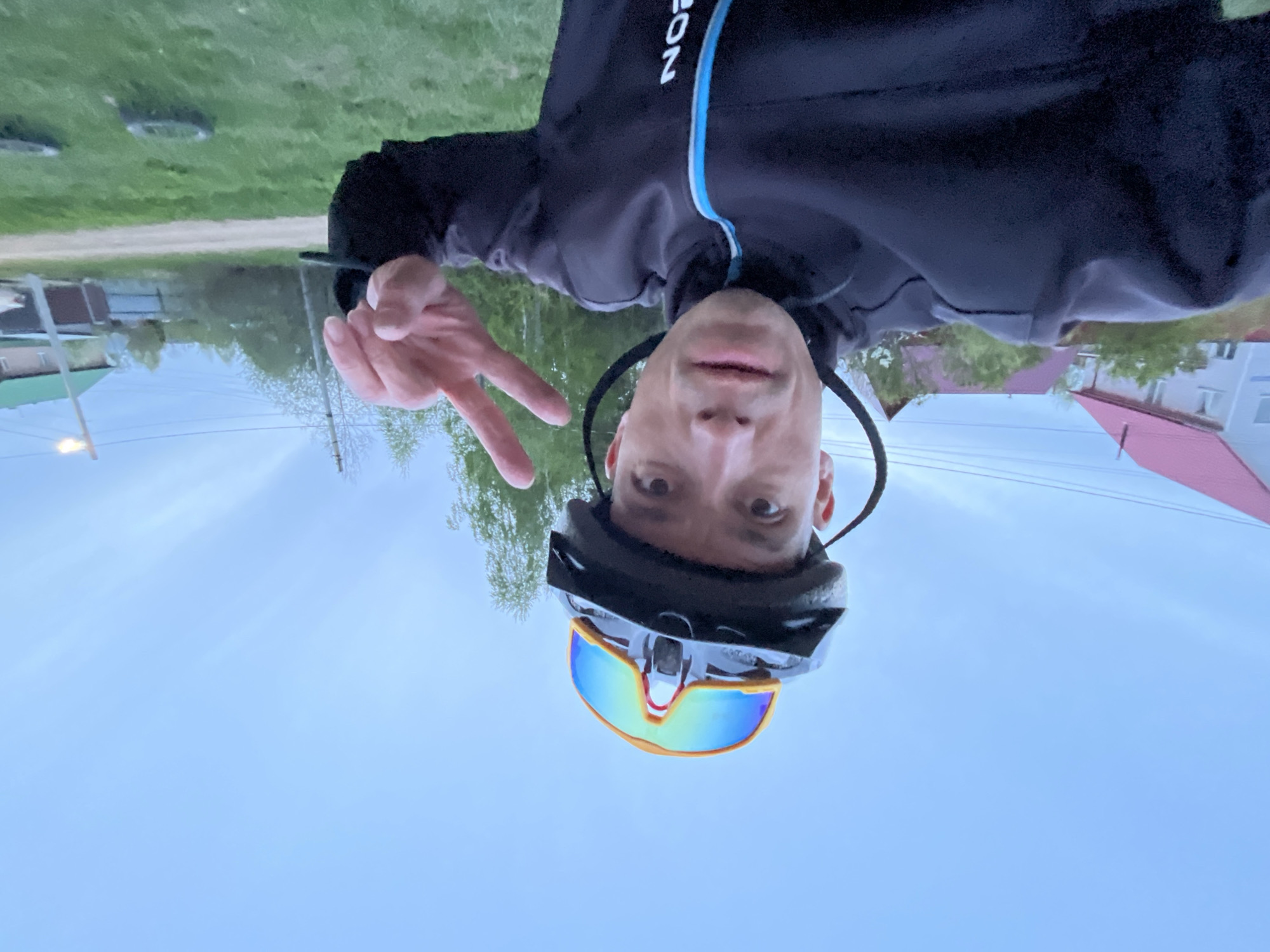 A man is photographed upside down, wearing a black cycling jacket with a blue stripe and black cycling helmet with reflective sunglasses. He is holding up two fingers in a peace sign. The backdrop includes a grassy area, a path, trees, houses with pitched roofs, and a light post against a cloudy sky. Based on the architectural style and general scenery, the location appears to be in a residential area, consistent with the city of Chaikovsky, Russia.