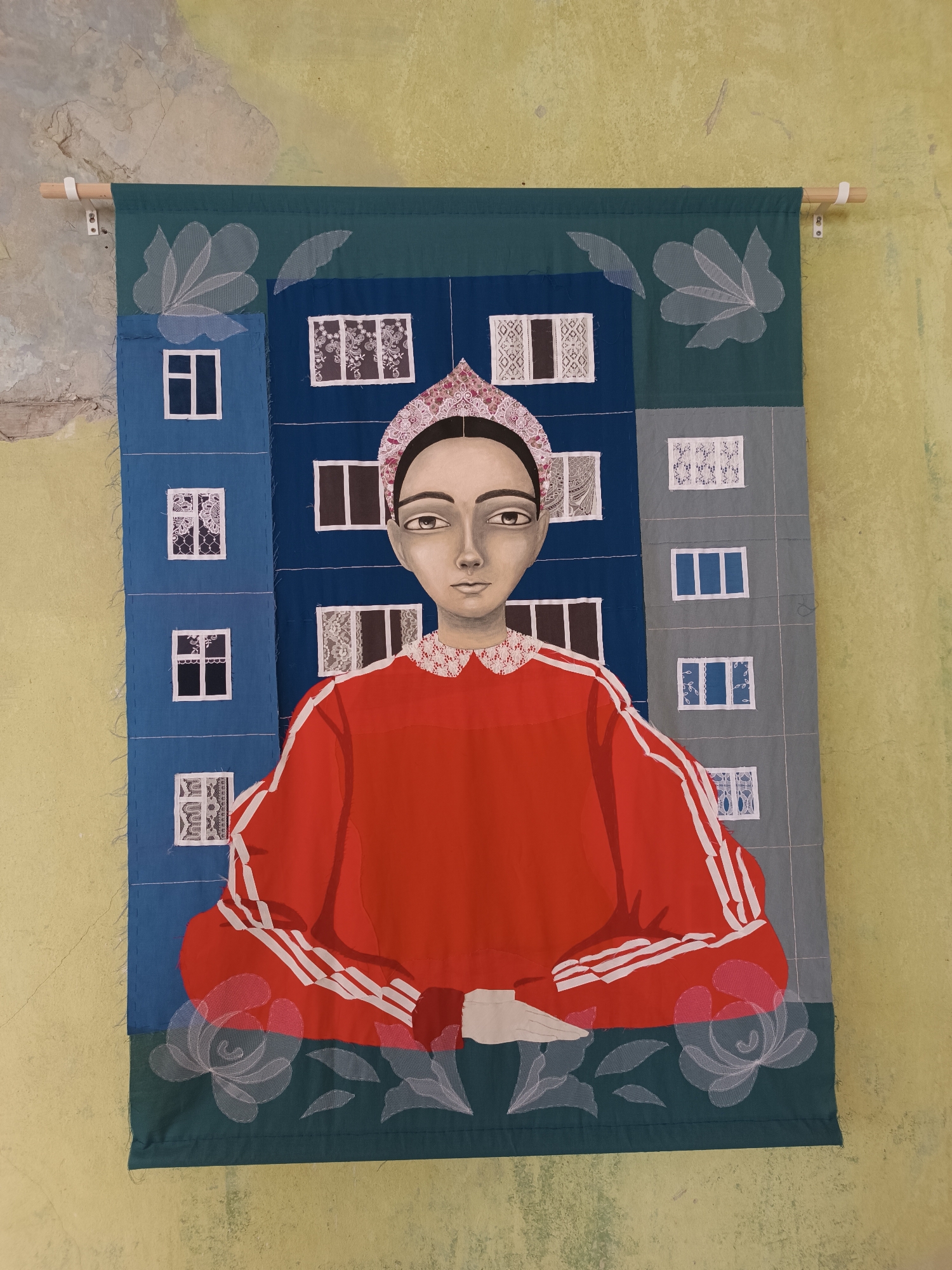 This is a photograph of a fabric tapestry hanging on a textured, yellow and grey wall, likely in an indoor setting. The tapestry depicts a stylized portrait of a young person against a backdrop of apartment building facades. The person is rendered in a naive, almost folk-art style, with large, dark eyes and a serene expression. They wear a red garment with white stripes on the sleeves, reminiscent of athletic wear, and a decorative headband. The background is composed of fabric squares representing blue and grey apartment buildings with various window designs, some featuring lace-like patterns. Floral motifs in a pale, translucent fabric are embroidered along the bottom edge of the tapestry. A wooden rod is attached to the top of the tapestry with white brackets, securing it to the wall. The overall impression is one of a piece of art displayed in a somewhat rough-hewn environment.