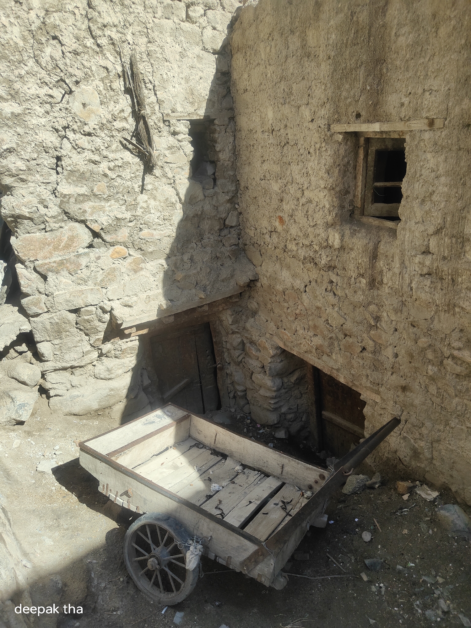 The image depicts a rustic scene likely in Leh, India, with a focus on weathered architecture and an old wooden cart. The dominant structures are made of rough, uneven stone and mud, suggesting an ancient or traditional construction style. A prominent feature is a small, dark opening in the wall, possibly a window or vent, above a wooden doorway set into an alcove.
In the foreground, a weathered wooden cart with two spoked wheels rests on the dusty ground. The cart's wooden planks are aged and worn, and its metal hardware shows signs of rust. The cart is positioned in a way that suggests it is no longer in active use, casting a distinct shadow on the ground.
The surrounding environment is characterized by debris and uneven terrain, consistent with an outdoor, perhaps rural or semi-abandoned, setting. Sunlight casts sharp shadows, highlighting the textures of the stonework and the cart. The overall impression is one of time-worn materials and a sense of history.