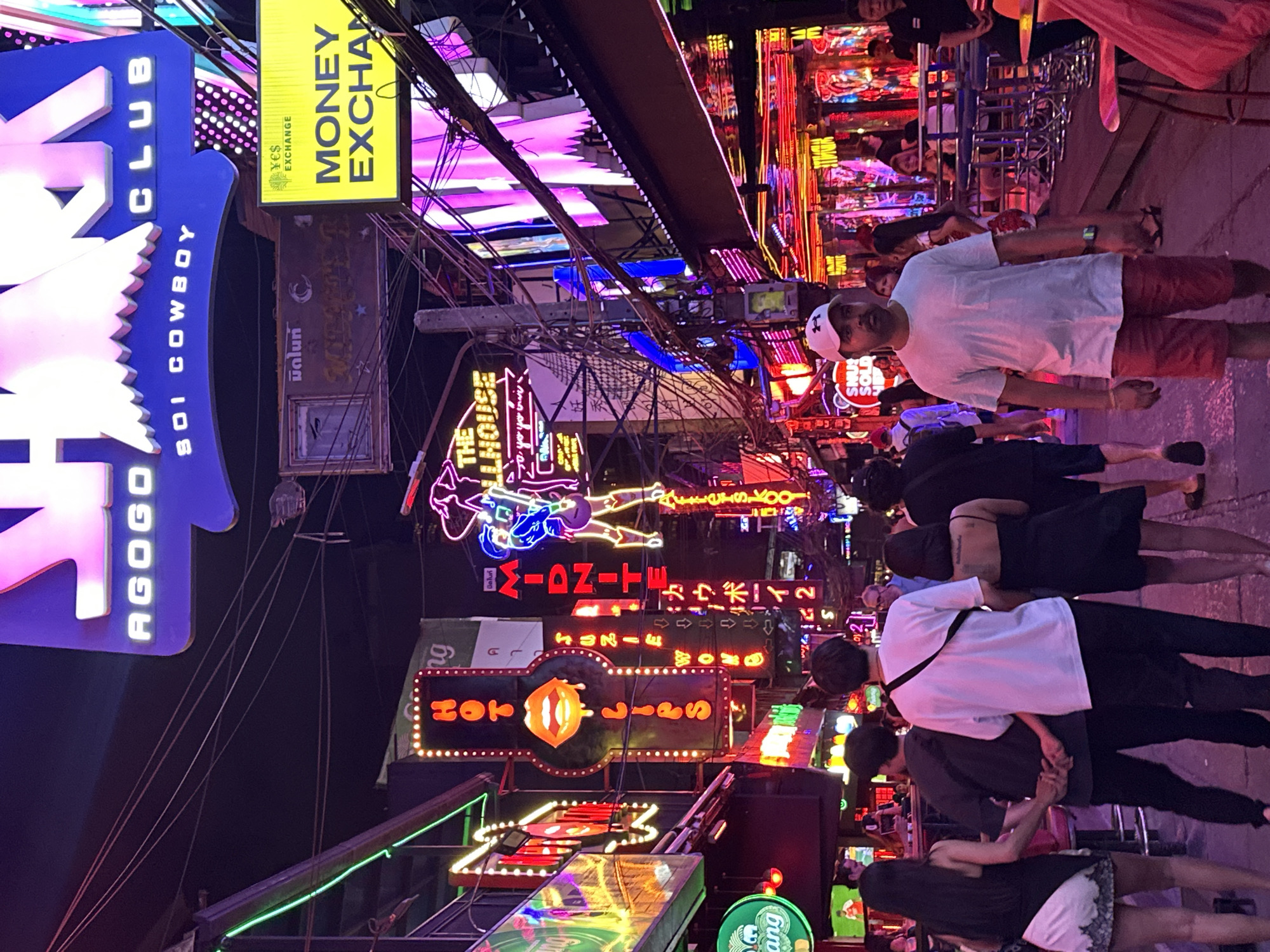 This photograph captures a street scene in Bangkok, Thailand, notable for its vibrant nightlife. The presence of numerous neon signs, including those for "Money Exchange," "AGOGO CLUB," "HOT LIPS," and "MIDNITE," indicate a commercial and entertainment district. People are visible on the sidewalks, walking along the street. A man wearing a white t-shirt and red shorts is seen on the right side of the frame, while a few young couples are seen walking in the middle. The signs "SOI COWBOY" and "THE ILLHOUSE" further identify the location. The urban environment is filled with overhead power lines and illuminated signage.