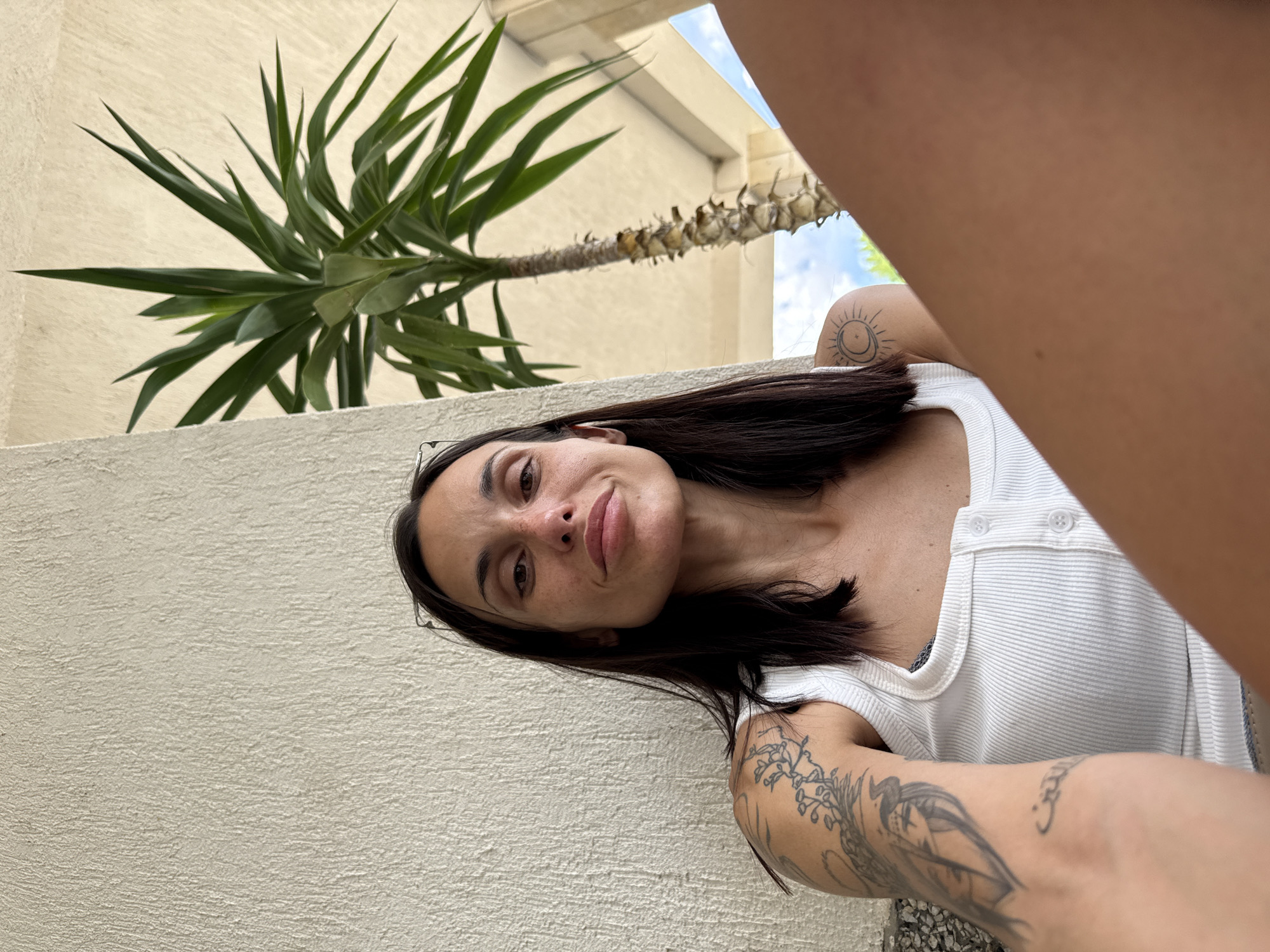 A woman is pictured seated outdoors, leaning against a textured white wall, with a palm tree behind her. She is looking towards the camera with a neutral expression. Her dark hair is shoulder-length, and she is wearing a white ribbed tank top. Tattoos are visible on her left arm and shoulder, including a sun and moon design. The environment suggests a warm climate.