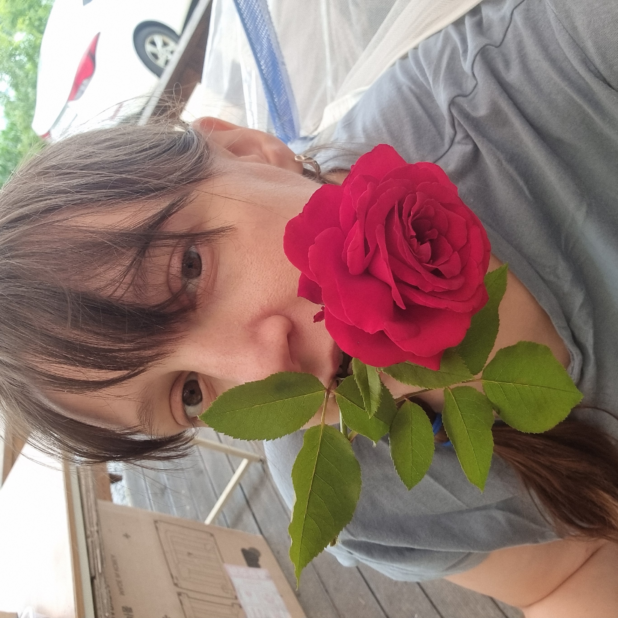 In this close-up, upside-down photograph, a person's face is partially obscured by a vibrant red rose held in front of it. The subject has dark brown hair that falls across their forehead and over their right eye. Their left eye, a deep brown, is visible, as are parts of their nose and cheek. A small, silver earring is visible on their left earlobe. The person is wearing a grey t-shirt, and the fabric is visible in the background, showing soft folds and texture.
The rose is the focal point, with its rich red petals tightly furled in the center and opening outwards. Several bright green leaves with serrated edges are attached to a stem extending from the rose, with one leaf positioned near the person's nose and mouth.
In the blurred background, a white vehicle is partially visible, with a black tire and wheel. Above the vehicle, some greenery suggests an outdoor setting, possibly with trees. The ground appears to be made of weathered wooden planks. The lighting suggests it is daytime, with natural light illuminating the scene. There is no visible text in the image.