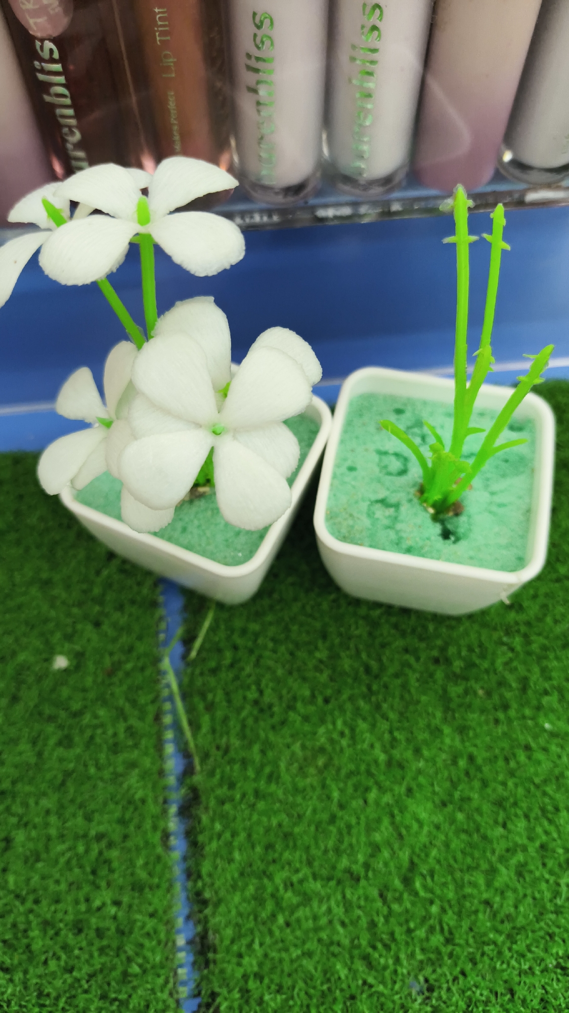 A close-up, vertical shot shows two small, square white pots containing artificial plants, placed on a textured green surface that resembles grass. The pot on the left holds a cluster of white, five-petaled flowers with green stems. The pot on the right contains a single green stem with small, undeveloped buds. In the background, out of focus, are several cylindrical objects, likely cosmetic products, with visible labels. The items appear to be arranged on a shelf. The overall impression is that of a retail display. The location is noted as South Jakarta, Indonesia.