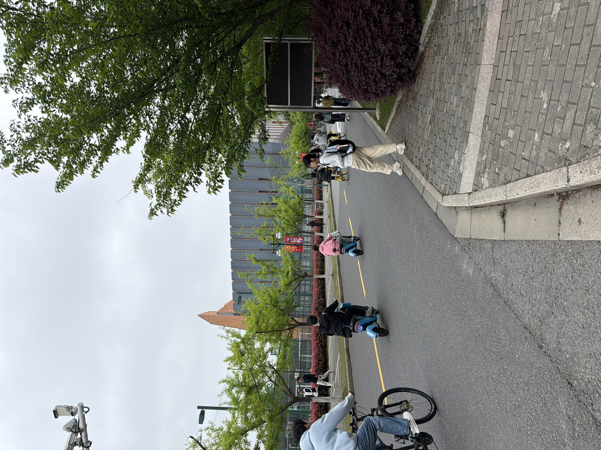 The image shows a street scene in the Jiangning District of China, likely on a university campus. The sky is overcast. A paved road runs through the scene with a yellow line indicating a lane. Several people are on the road, including one person riding a bicycle in the foreground. Further up the road, several people are riding small blue electric scooters. Two individuals are walking alongside the road on a paved area. Buildings with modern architecture are visible along the side of the road, with greenery including trees and bushes. A signpost is next to the paved walkway.