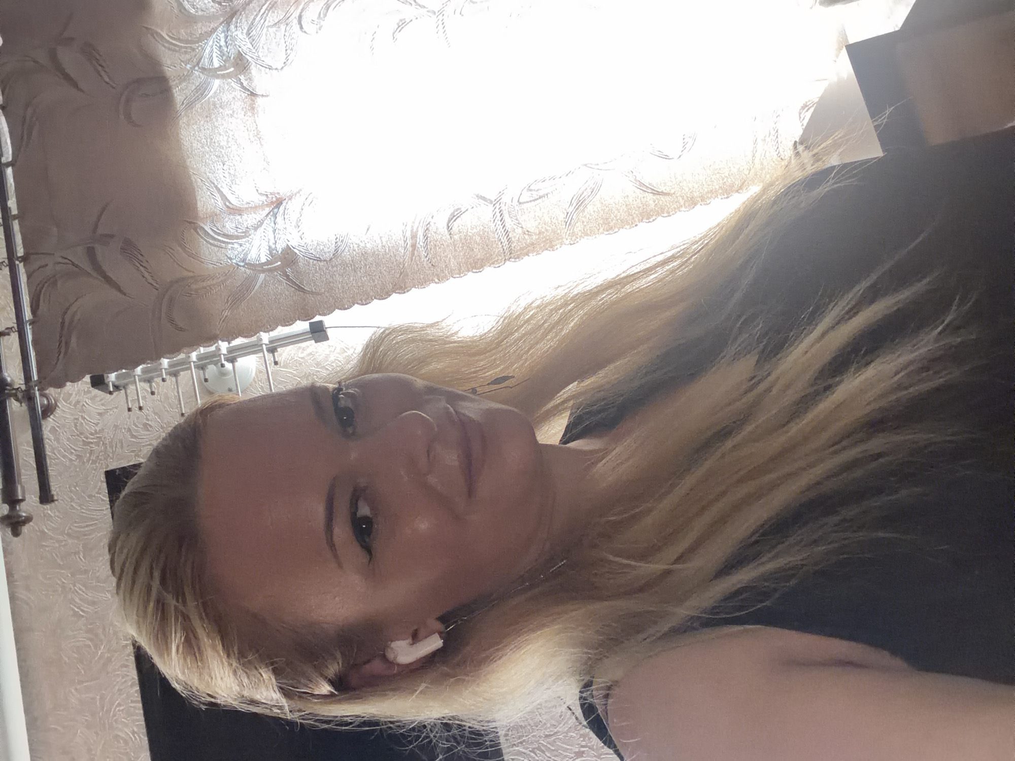 A fair-skinned woman with blonde hair is pictured in a close-up selfie. She is wearing a black top and white earbuds. Her hair is styled down and slightly tousled. The background appears to be an indoor setting with light-colored, textured wallpaper and sheer curtains that are lit from behind, creating a bright, hazy effect. The lighting suggests it is daytime.