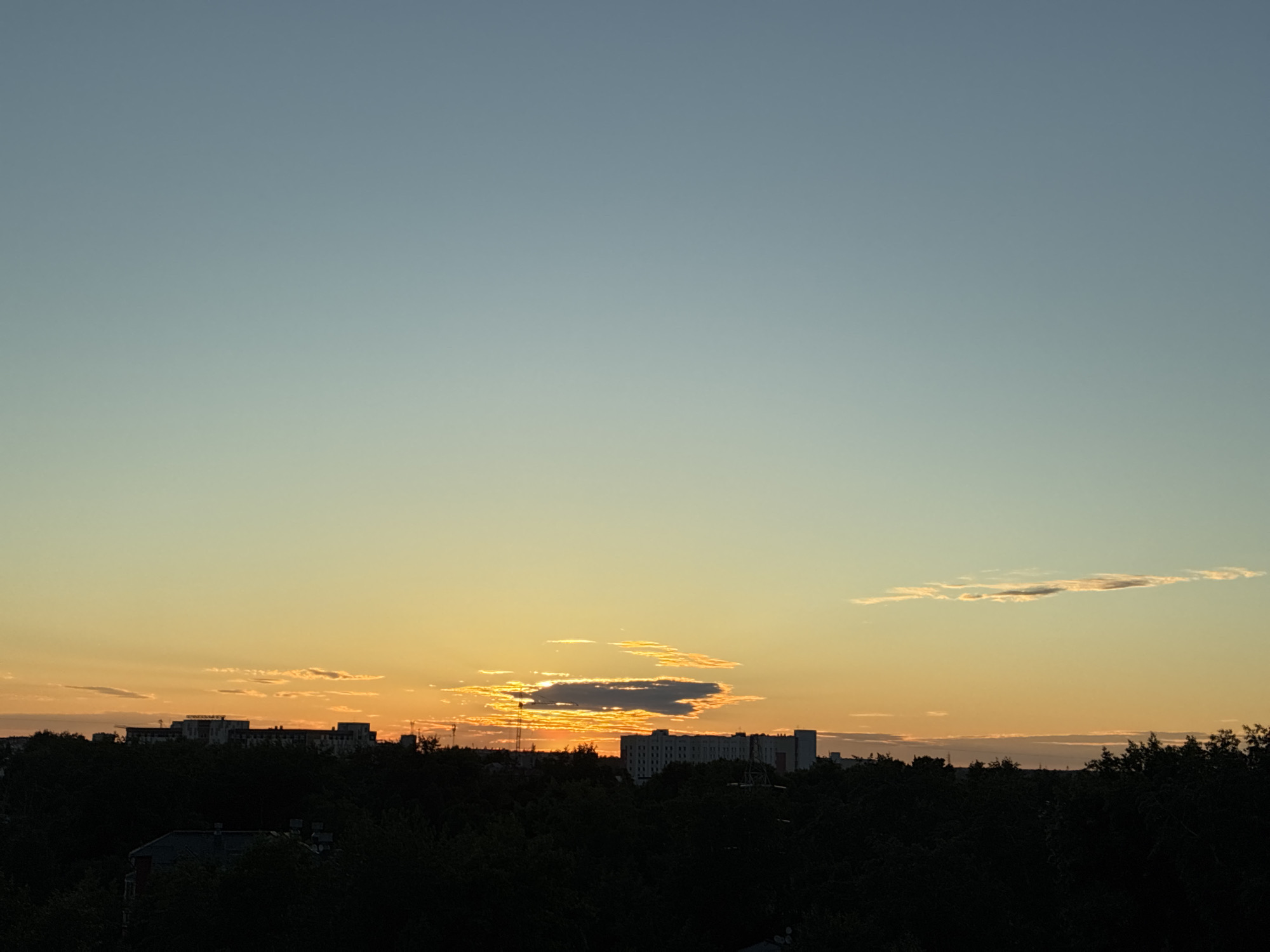 A wide shot captures a city skyline at dusk, with the sky transitioning from a pale blue at the top to warm orange and yellow hues near the horizon. Silhouetted against this vibrant sky is a dense line of dark green trees, interspersed with the outlines of buildings and what appear to be radio towers. Several multi-story buildings are visible, with one prominent structure featuring a sign that is unreadable due to the distance and lighting. The light creates a dramatic contrast between the illuminated sky and the darkened landscape.