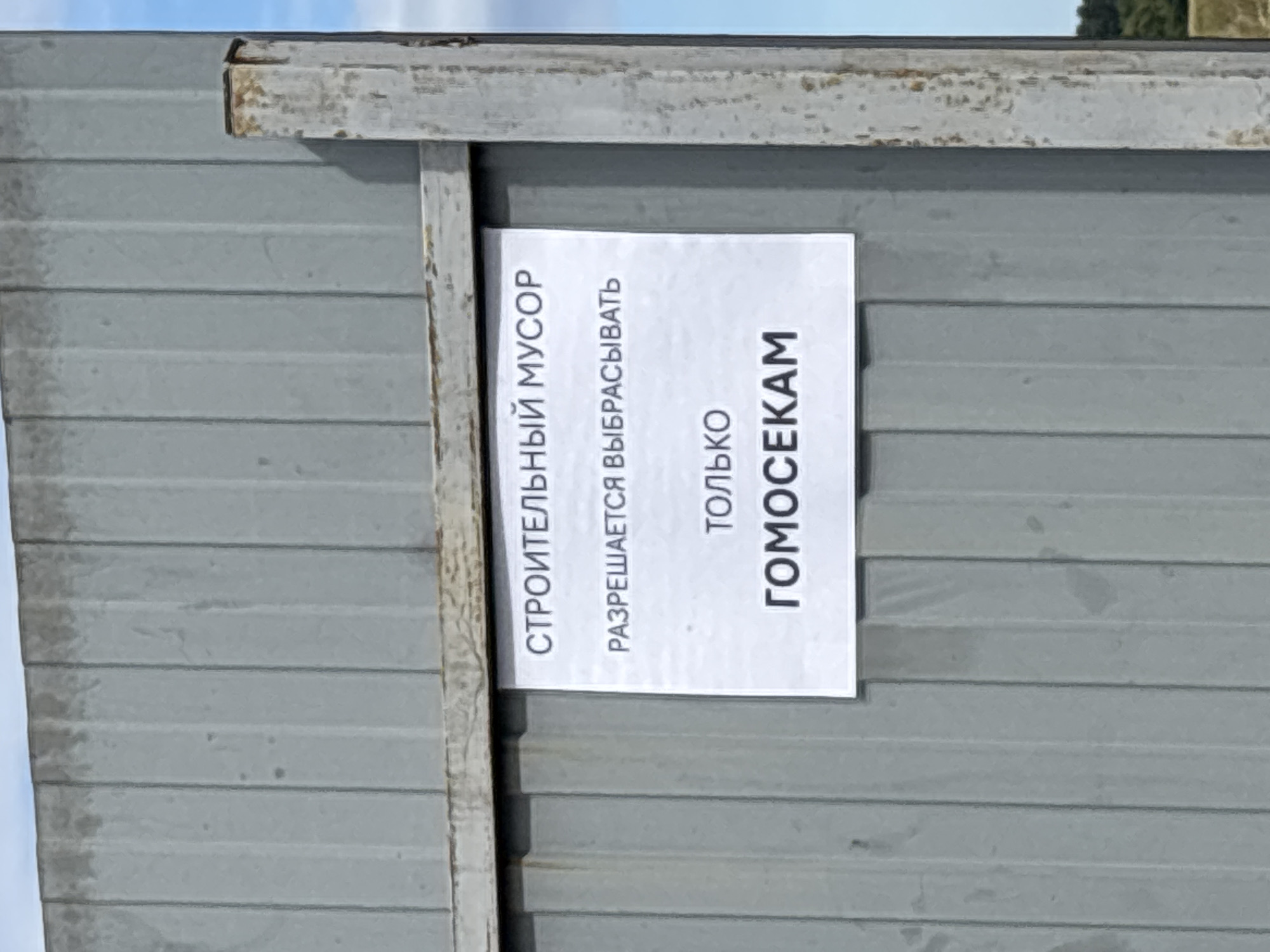 A sign is affixed to a weathered gray metal fence. The sign, printed in Russian, reads "CONSTRUCTION WASTE. ONLY GOMOSEKAM ARE ALLOWED TO DISPOSE OF IT." The fence appears to be located in an outdoor environment, possibly a construction site or waste disposal area. The background shows a blue sky and some greenery, suggesting an urban or suburban setting. The overall condition of the fence and sign indicates some age and exposure to the elements.