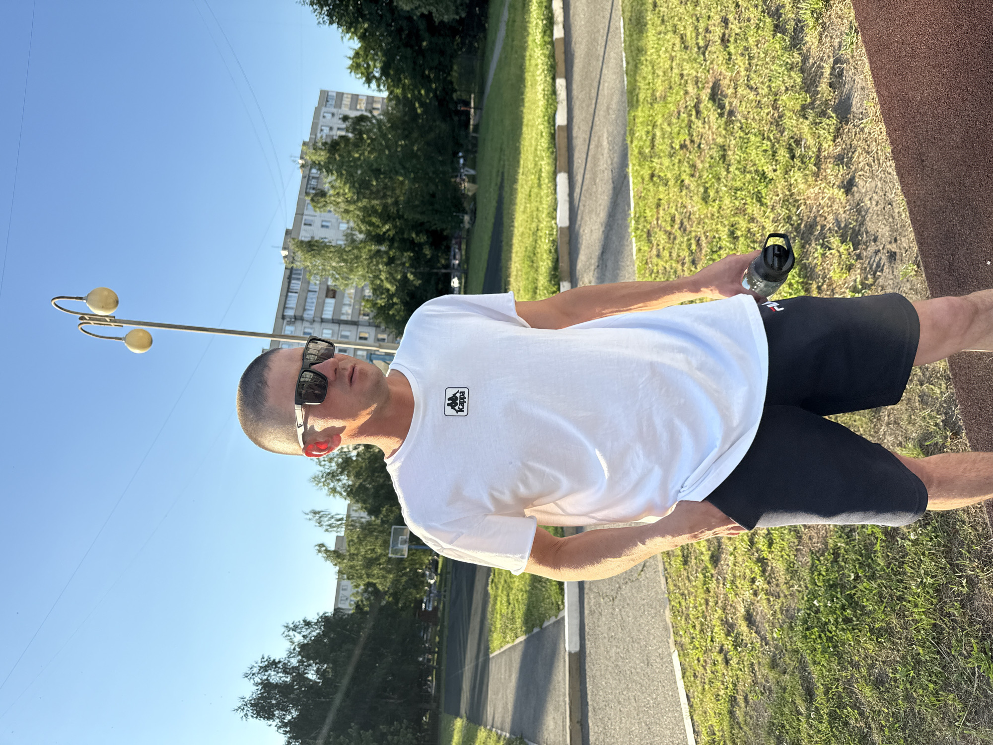 A man wearing sunglasses, a white t-shirt with a Kappa logo, and dark shorts stands in a park with a basketball court and trees. A building and a lamppost are visible in the background. He is holding a black water bottle. This scene is located in Stary Oskol, Russia.