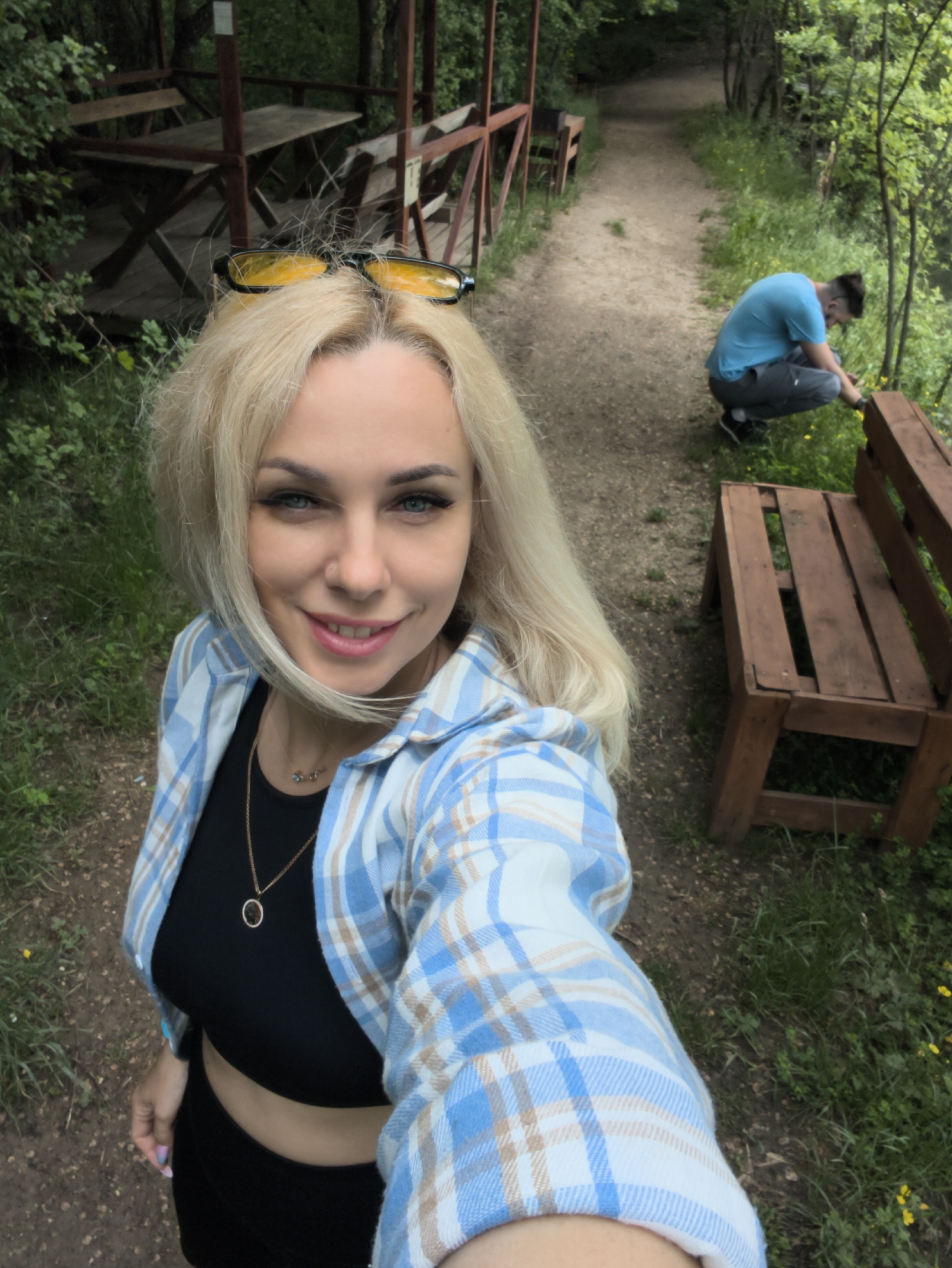 A medium, angled shot shows a blonde woman holding the camera taking a selfie outdoors. She wears black sunglasses on her head, a gold chain necklace, a black top, and a blue plaid shirt. Behind her is a dirt path with a wooden bench and a man wearing a blue shirt and gray pants squatting and picking flowers. To the left, there is a wooden gazebo. The location appears to be in a park or wooded area in Терновский округ, Ukraine.