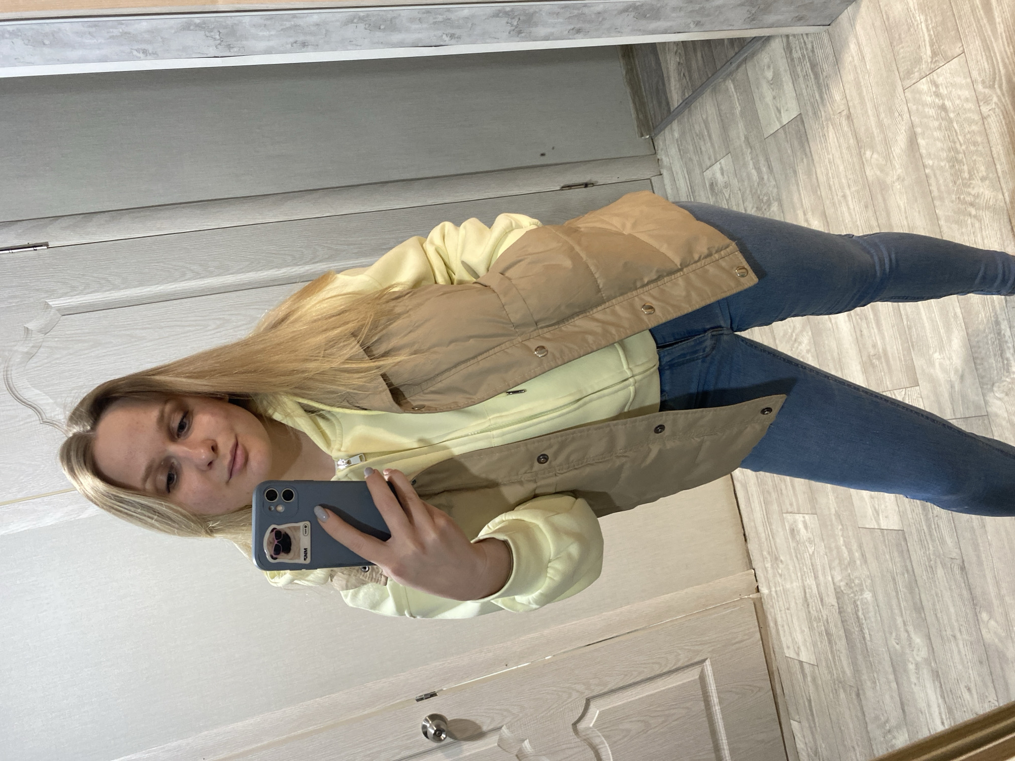 A young woman with blonde hair takes a mirror selfie indoors. She is wearing a light yellow hooded sweatshirt, a tan puffer vest, and blue jeans. Her phone, decorated with a sticker of a pug wearing sunglasses, is visible in her left hand. The background shows a light-colored door with a silver knob on the left and a wall with a mirror on the right. The floor is a light-colored wood laminate. The scene appears to be within a residential dwelling.
