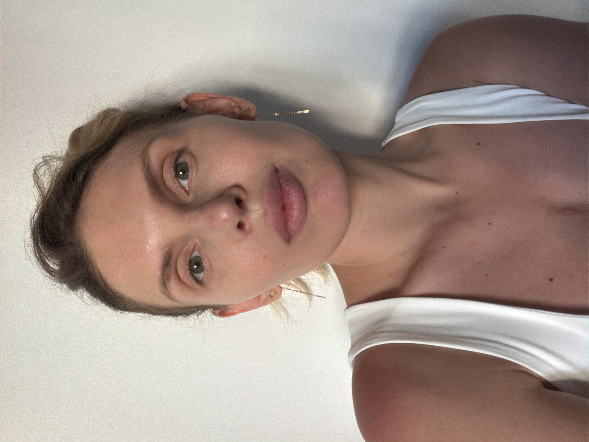 A medium shot depicts a fair-skinned woman with blonde hair, wearing a white tank top. She has light green eyes and her hair is partially pulled back. She is wearing a long, thin gold earring. The background is a plain, light-colored wall. No location can be determined.