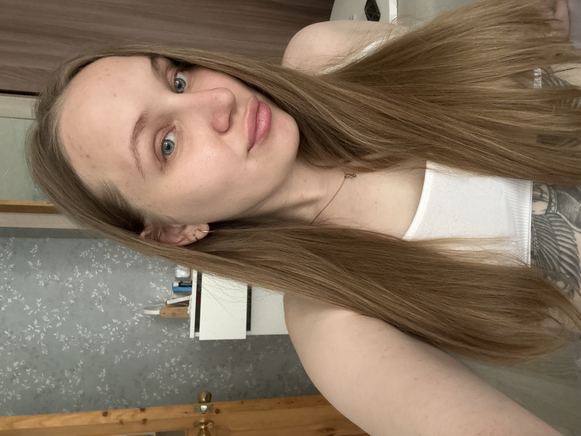 A young, light-skinned woman with long, light brown hair, blue eyes, and a fair complexion poses for a selfie. She wears a white tank top. A portion of a tattoo is visible on her upper chest/shoulder. She is indoors, positioned in front of a wall with floral wallpaper and a wooden dresser or headboard. A partially visible white cabinet or shelf unit is in the background. There's no indication of Chaikovsky, Russia.