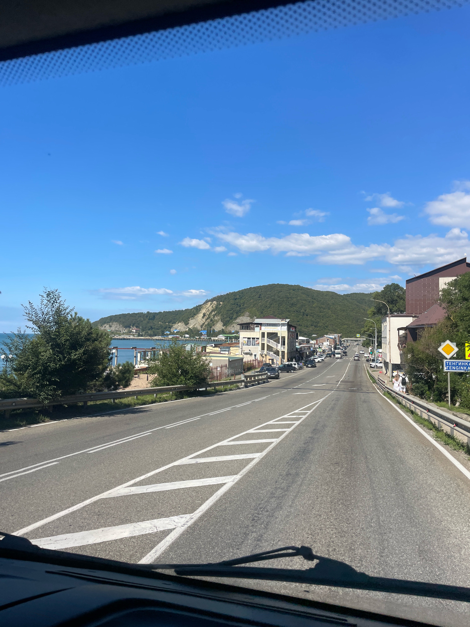 The image captures a view from a vehicle looking down a road that curves to the left, with mountains in the background and the sea visible on the left. The road is paved and has clear lane markings. Buildings and trees line the right side of the road, and a sign with Russian text and "TENGINKA" is visible. Several cars are parked or driving on the road, and two people are walking on the sidewalk. The sky is blue with scattered white clouds. The overall scene depicts a coastal town or village.
