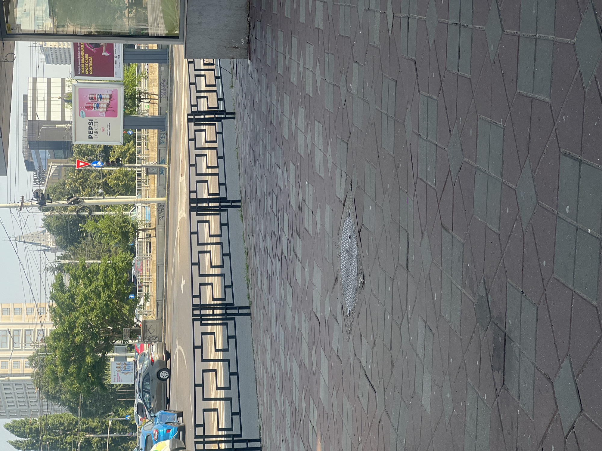 The image shows a street scene in Iasi, Romania, with a view of a sidewalk, a street with cars, and buildings in the background. A black metal fence with a geometric pattern runs horizontally across the middle of the image, separating the sidewalk from the street. The sidewalk is paved with interlocking geometric tiles in shades of red and grey, some of which are uneven or broken. A manhole cover is visible on the sidewalk.
In the street, several cars are visible, including a blue car and a grey sedan. There is a traffic light and a pedestrian crossing. In the background, there are trees and several buildings, including a modern office building with a glass facade and another building with a classical architectural style. Billboards are also visible, one of which advertises Pepsi. The sky is clear and appears to be daytime. The overall impression is an urban environment on a sunny day.