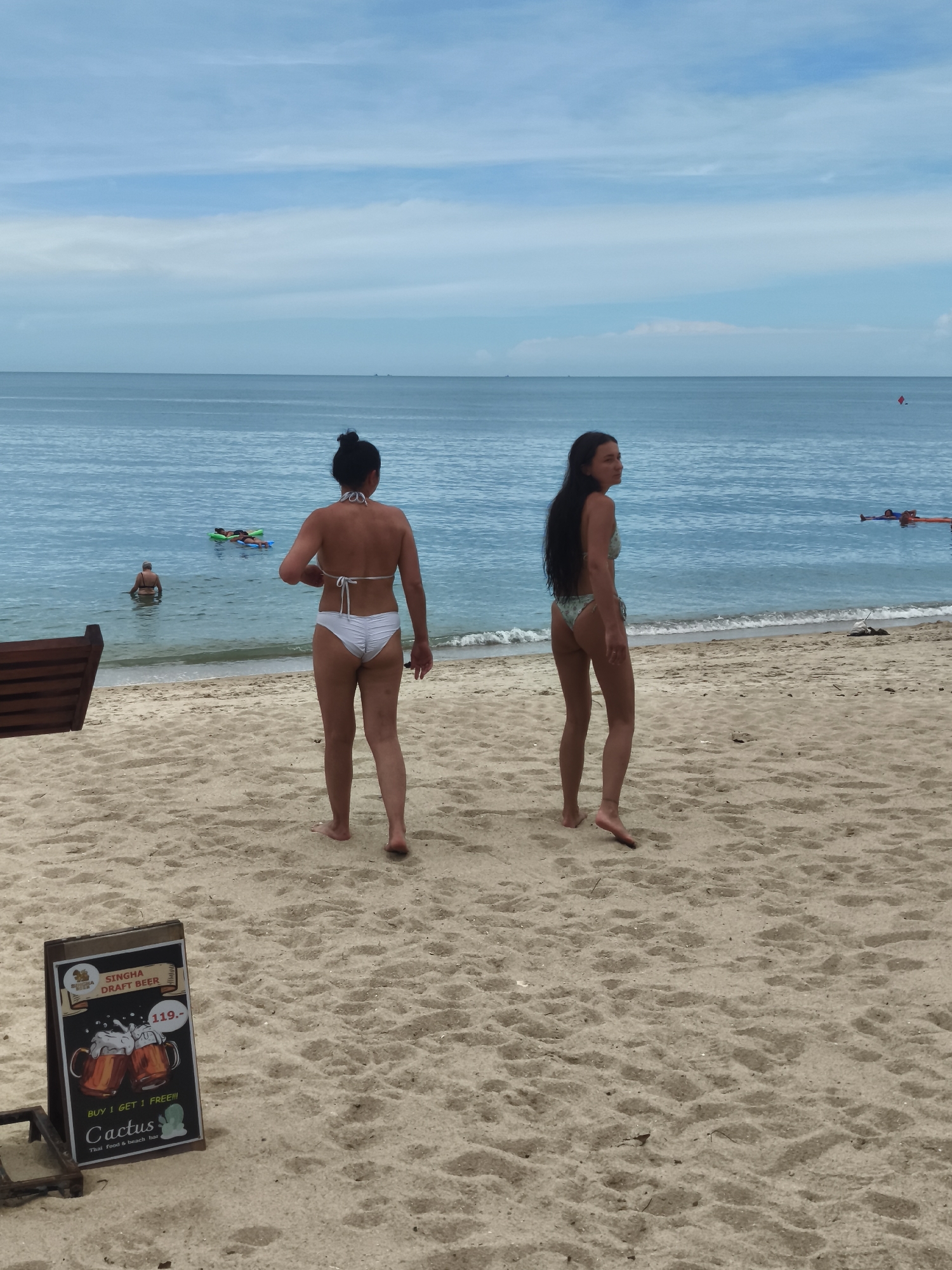 At Baan Lamai beach in Thailand, two women in bikinis are standing on the sand facing the ocean. The woman on the left has her dark hair pulled up in a bun and is wearing a white bikini. The woman on the right has long, dark hair and is wearing a patterned bikini. In the shallow water, another person is visible, and further out, two people are lying on inflatable rafts. The sky is cloudy with a calm, blue sea. In the foreground, a small A-frame sign advertises "Singha Draft Beer" with a "Buy 1 Get 1 Free!!!" offer and lists "Cactus Thai Food & Beach Bar". A portion of a wooden beach chair is visible on the left side of the frame.