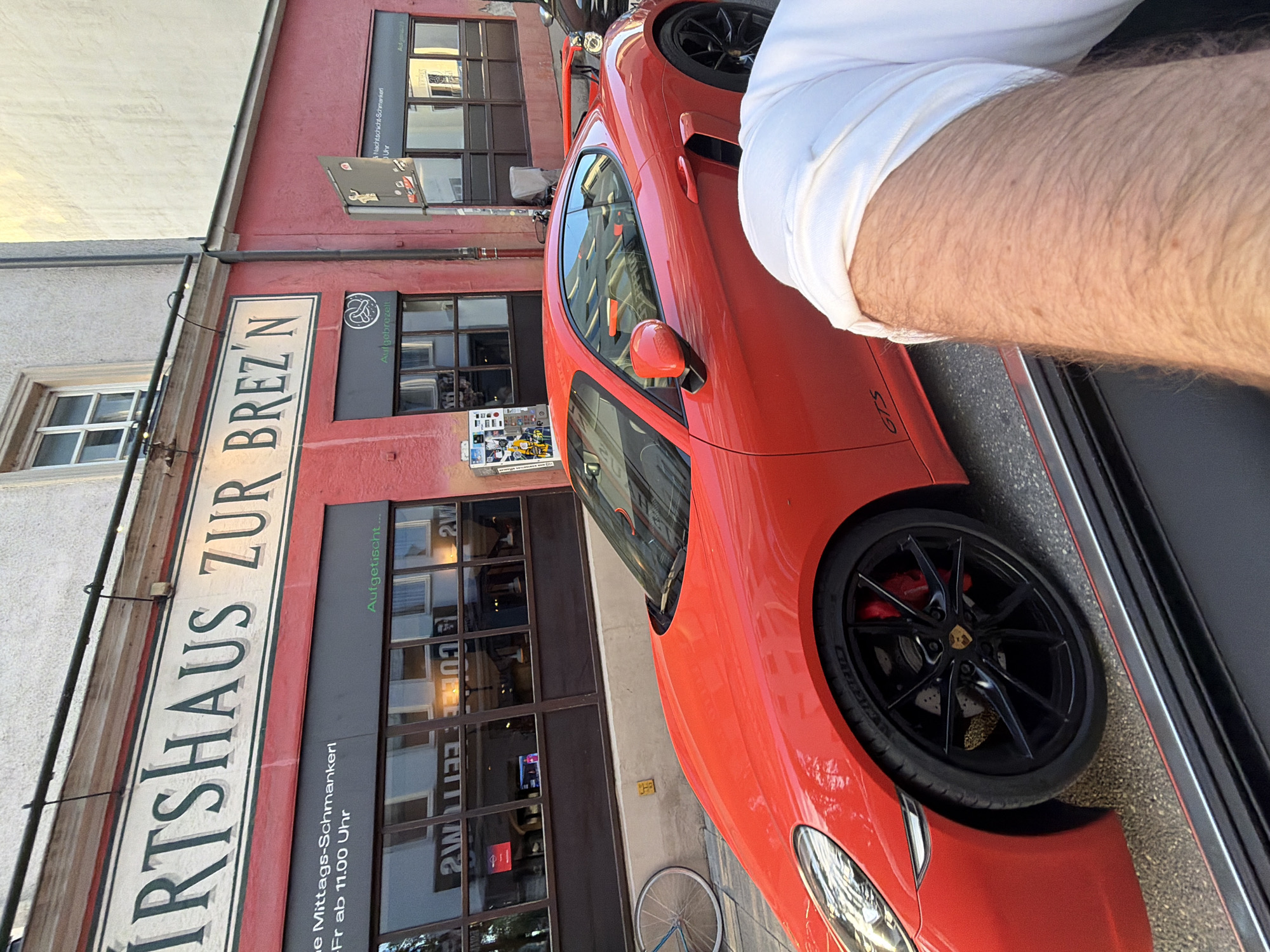 The image shows a red Porsche sports car parked on a street in Munich, Germany. The car is in the foreground, and a building with a sign that reads "WIRTSHAUS ZUR BREZ'N" is in the background. The building appears to be a restaurant or pub. There is a person's arm, wearing a white shirt, visible in the lower right corner of the image, suggesting the photo was taken from inside another vehicle. The street is paved, and there is a bicycle leaning against a wall on the left side of the image. The overall lighting suggests it is daytime, with sunlight casting shadows. The architecture and signage are typical of a European city.