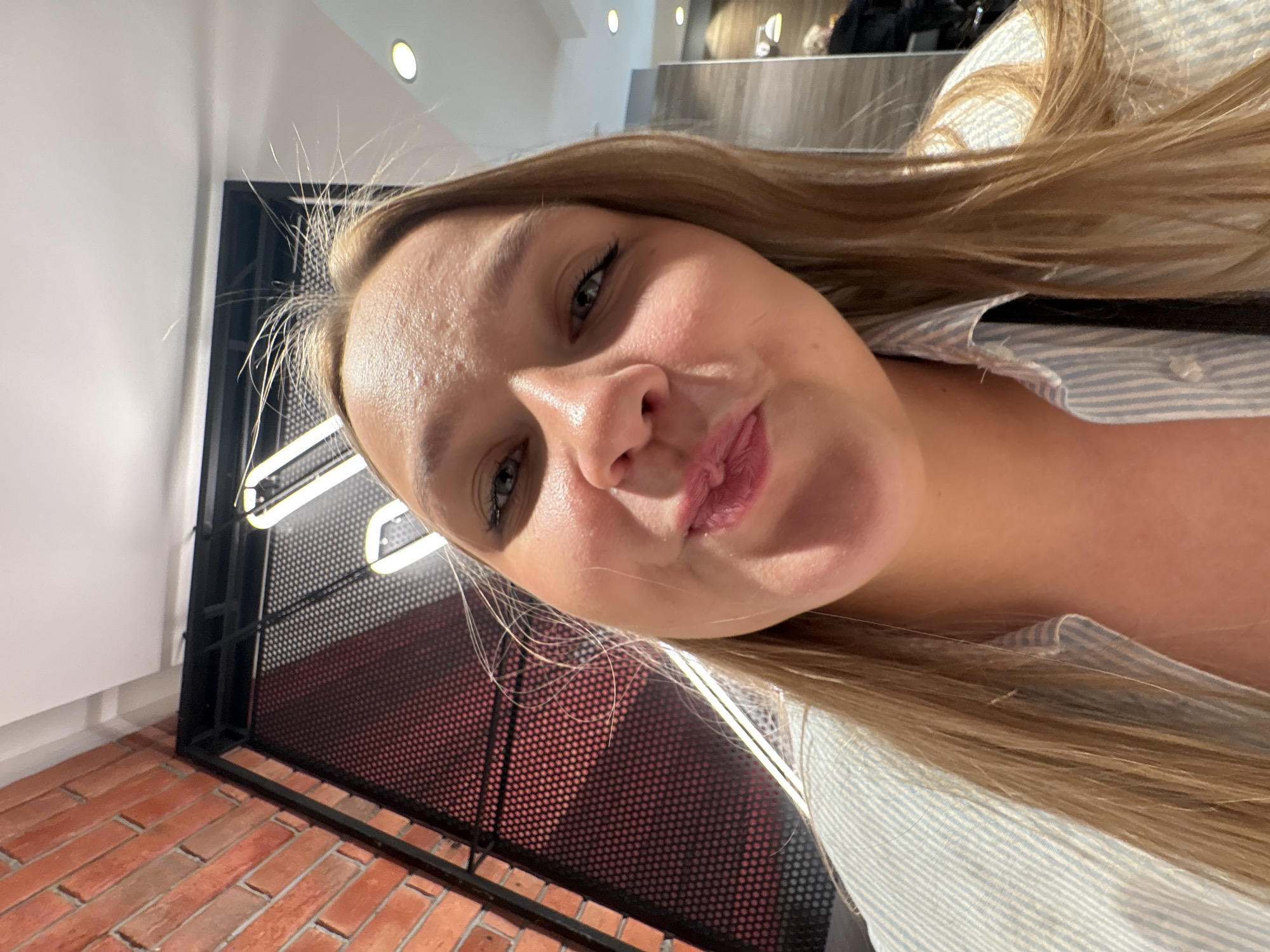 A selfie shows a woman with blonde hair and light skin pursing her lips. She is wearing a light blue and white striped shirt. Behind her is a black metal grid with a lit neon sign. In the lower-left corner, there is a brick wall. The location is Izhevsk, Russia.