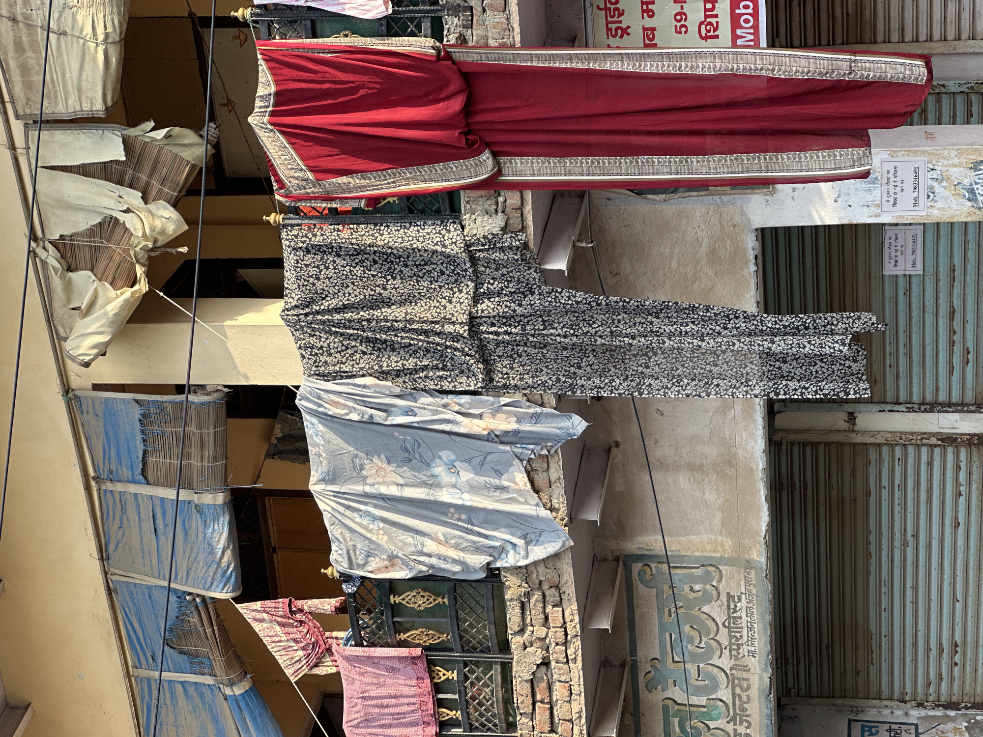 The image depicts an outdoor urban scene in Mathura, India, characterized by the presence of various textiles hung on a wire between buildings. At the top, a red garment with intricate borders is visible, followed by a black and white floral patterned fabric, and a blue floral patterned sheet hanging below. These items appear to be drying or displayed outdoors.
The buildings framing the scene showcase weathered facades. One has metal shutters and signs with Hindi script, including the name “निरन्जन लाल अर्जुन पुरा मधुर” and the phrase “ये दुकान चौराह पर शिफ्ट हो गई है परिक्रमा मार्ग पर" along with a mobile number (7983316491).
Above the textiles are makeshift awnings or shades made of tattered fabric and bamboo, attached to the buildings. The environment appears cluttered with wires running across the scene and aged brickwork.