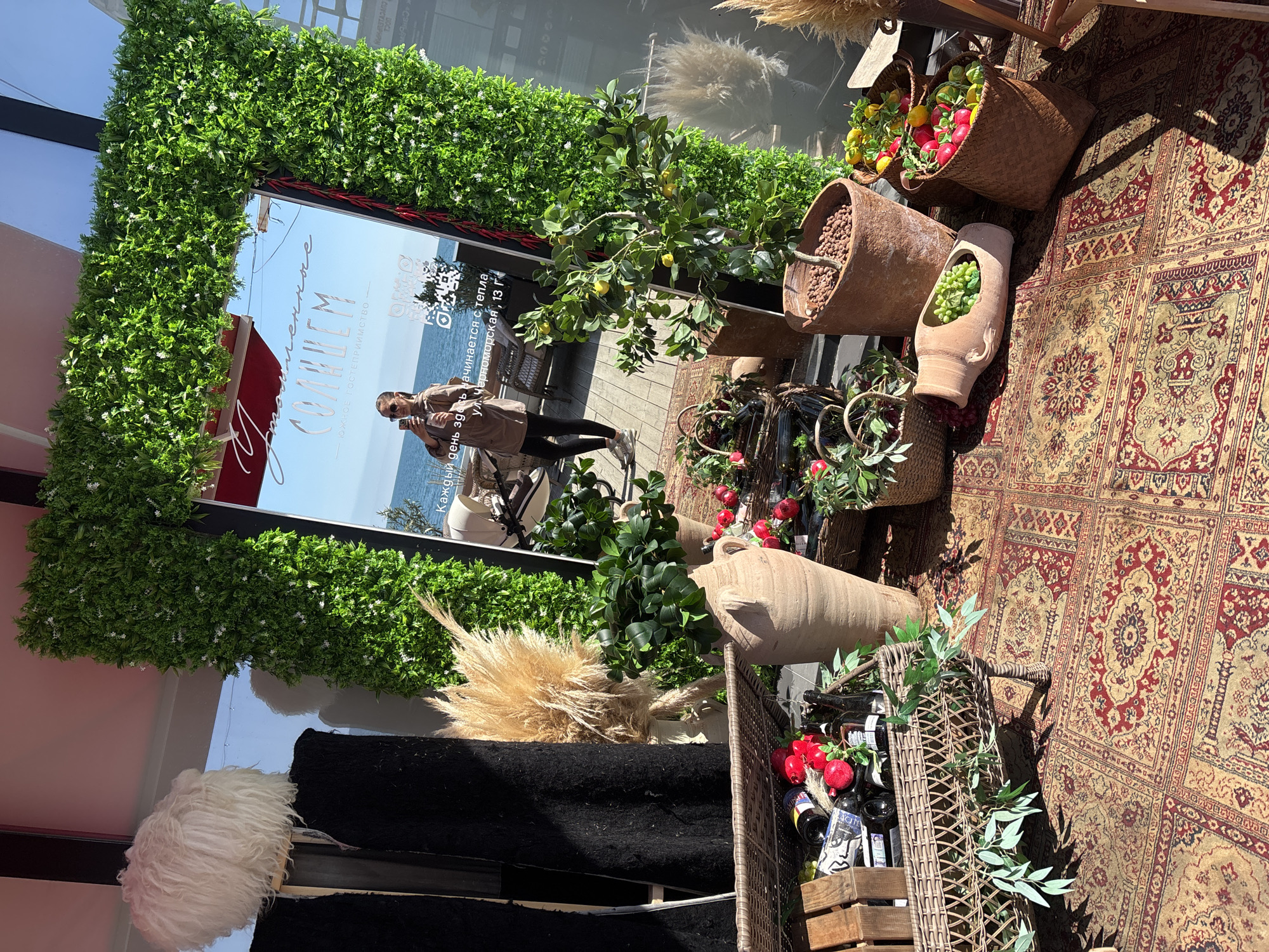 The image shows an outdoor decorative setup, presumably for a restaurant or business in Sochi, Russia, located at 13 Chernomorskaya Street. A large mirror with an artificial green frame reflects a view of the sea and a woman taking a picture. Various baskets and clay pots containing fruits, vegetables, and plants are arranged on a patterned rug. A wooden crate holds bottles and red fruits. Overall, the scene suggests a Mediterranean or coastal theme.