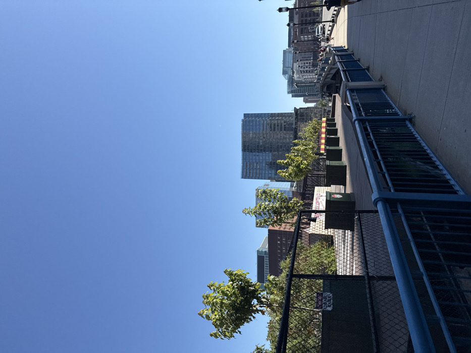 This image displays an urban landscape in Boston, Massachusetts, specifically a view from a bridge over the Fort Point Channel looking towards the Seaport District. The scene is illuminated by bright daylight under a clear, cloudless blue sky.
In the foreground, a dark blue metal railing parallels a concrete pedestrian walkway, curving gently to the right. To the railing's immediate left, a black chain-link fence encloses an area containing green foliage and obscured structures, with a white sign bearing red text, "DO NOT ENTER," and a smaller "DANGER" warning. A linear arrangement of small, green-leafed trees in dark square planters runs along the left side of the walkway, extending into the midground. One planter or an adjacent structure displays a red-and-white sign partially legible as "LOBSTER & CO."
The midground features the continuing concrete walkway on the right, where multiple indistinct human figures are walking away from the viewer. Adjacent to the walkway on the right is a roadway populated by several white vehicles and additional pedestrians. Black lampposts line this roadway. The structural elements of the bridge, including concrete and metal, are visible below the railing and alongside the road.
The background is dominated by the modern skyline of the Seaport District, characterized by several tall, glass-faced skyscrapers that reflect the sunlight. A prominent, dark-paned glass building stands near the center. Additional brick and glass buildings complete the cityscape. The upper half of the image consists entirely of a clear blue sky. A solitary person, partially visible on the far right of the frame, walks away from the camera, wearing a dark shirt and light-colored shorts.