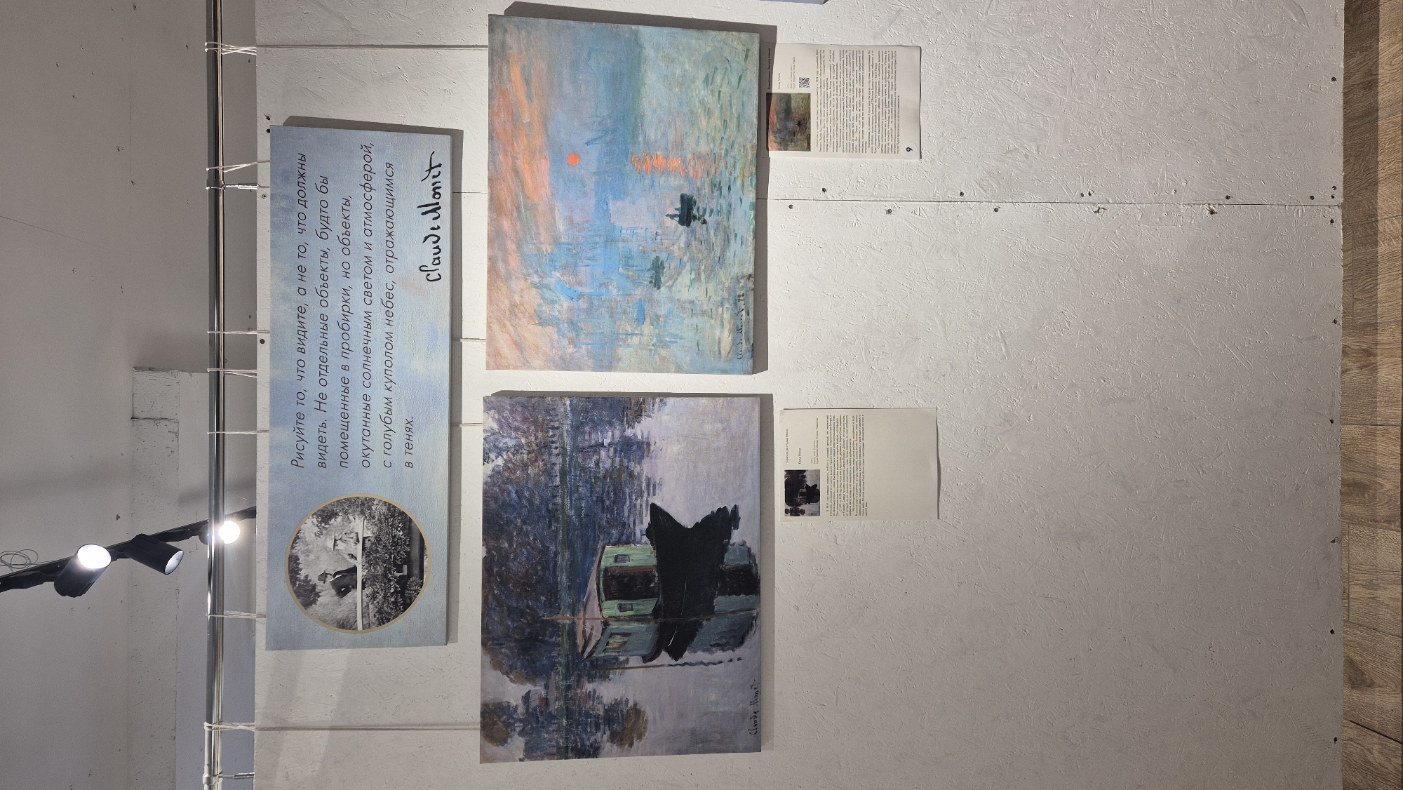 The image shows an art exhibition in Perm, Russia, likely at the Perm State Art Gallery, judging by the context of similar exhibitions. The exhibit features paintings by Claude Monet. In the center of the image, a large text panel with a circular image of two people standing near a bridge is displayed. The text on the panel is in Russian and quotes Monet: "Paint what you see, not what you think you should see. Not separate objects, but objects enveloped in sunlight and atmosphere, with a blue dome of heaven, reflected in the shadows." Below the panel, two Monet paintings are hung. The painting on the left depicts a boat on a body of water with trees in the background. The painting on the right is a landscape with a prominent red sun setting over a body of water with boats. Below each painting, there are informational plaques with smaller images. The wall the art is displayed on is a textured, light-colored surface. Track lighting is visible above the art. The floor is made of wood planks.