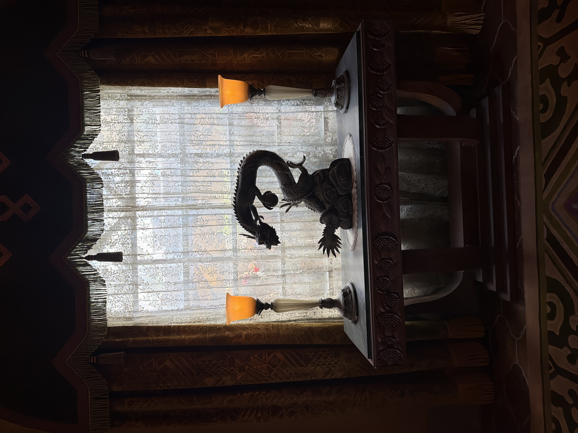 The image depicts an interior scene, likely in Chessy, France, featuring a window with ornate curtains and a carved wooden table. A dragon sculpture is prominently displayed on the table, positioned in front of the window, which is covered by a sheer, patterned fabric. Two lamps with orange glass shades are mounted on the table, one on each side of the dragon sculpture. The curtains are heavy and dark, with decorative fringe and tassels, framing the window. The overall aesthetic suggests a dramatic or theatrical setting, possibly a hotel lobby or a themed room. The lighting is dim, with the lamps providing a warm, ambient glow, and the sunlight filtering through the window creates a contrast between the illuminated background and the darker foreground. There is no visible text in the image.