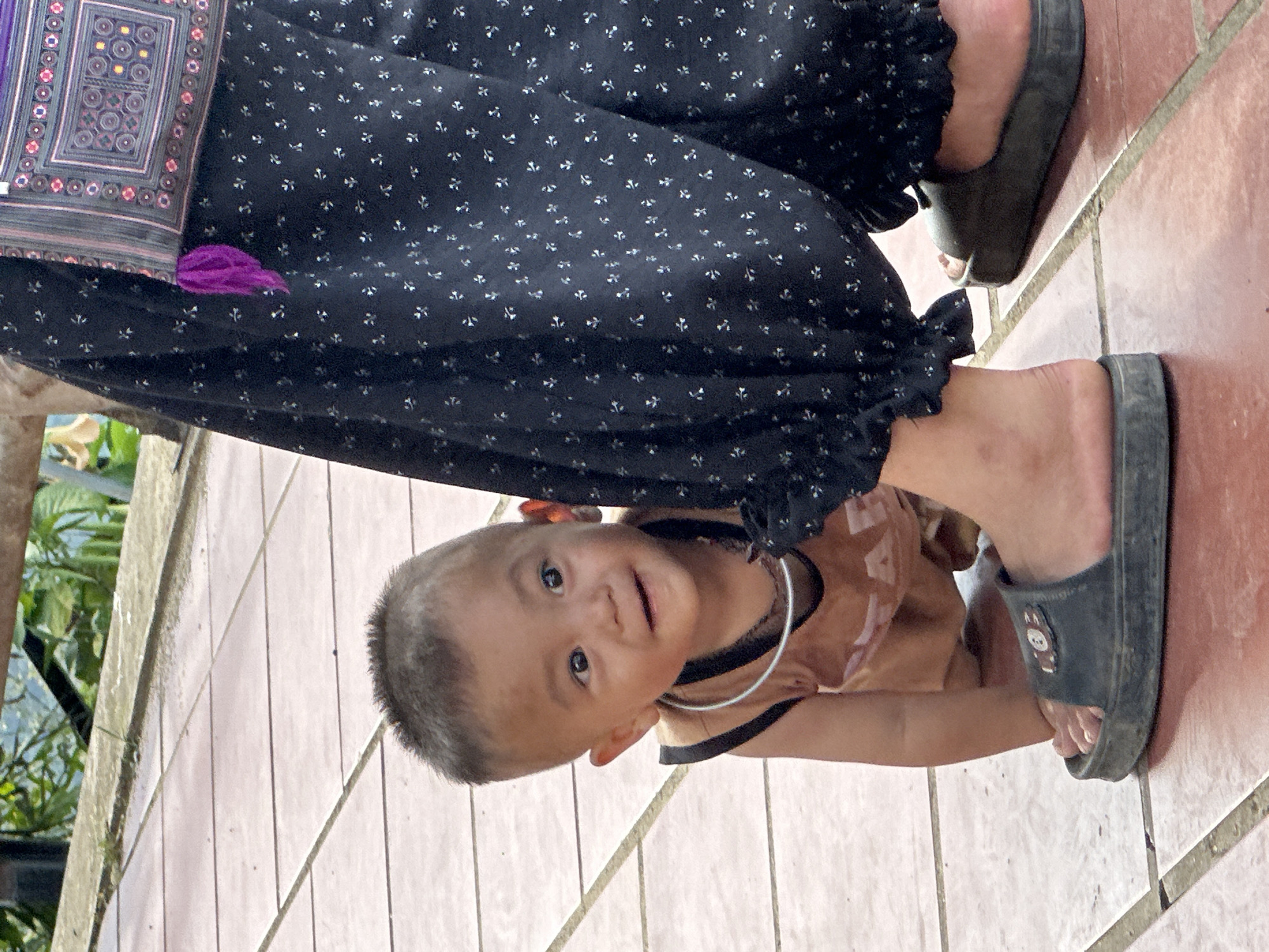 In Tu Le, Vietnam, a young child, approximately 1-2 years old, is captured in a crawling or crouching position, peeking from behind an adult's left leg. The child has short, dark hair, tan skin, and wears a sleeveless, light brown top with a dark trim. A thin, silvery-white metal ring rests around the child's neck. The child looks directly at the viewer with a curious, open-mouthed smile. The adult's lower legs are clad in dark, patterned trousers or a long skirt featuring small, light-colored repeating motifs, with ruffled hems above the ankles. The adult wears dark, flat slide sandals; the left sandal has a small, light-colored emblem. A patterned, woven bag with intricate geometric designs in red, purple, black, white, and yellow/gold, accented by a purple tassel, hangs from the adult's hip. The subjects are standing on a reddish-brown tiled surface with lighter grout lines. In the blurred background, green foliage and a light-colored concrete ledge are visible, suggesting an outdoor setting.