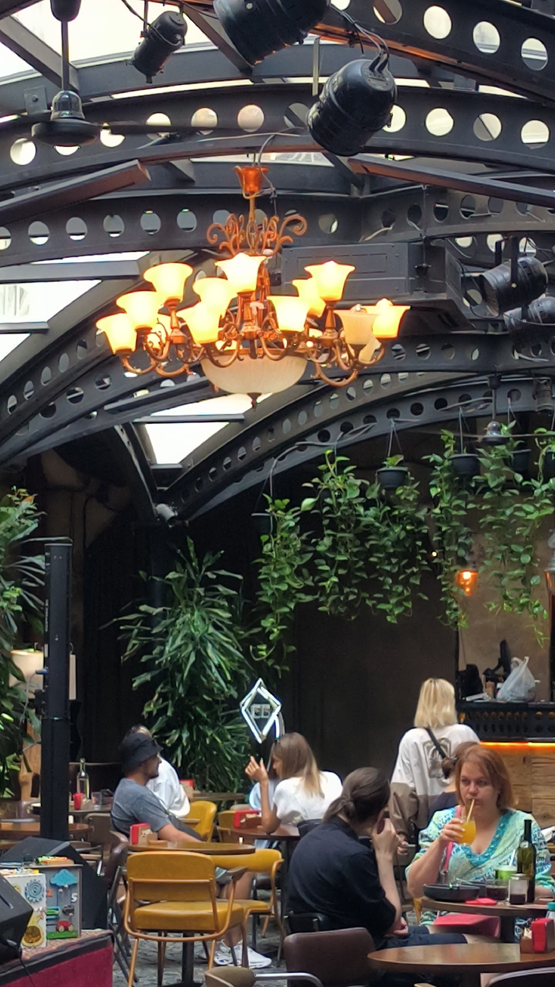 The image shows an indoor outdoor space that appears to be a restaurant or bar in Lviv, Ukraine. Several people are seated at tables, some of whom are interacting with each other, while others are drinking. There is a chandelier hanging from the ceiling and plants are visible throughout the space. The space has a modern industrial design with exposed metal beams and dark wooden accents.