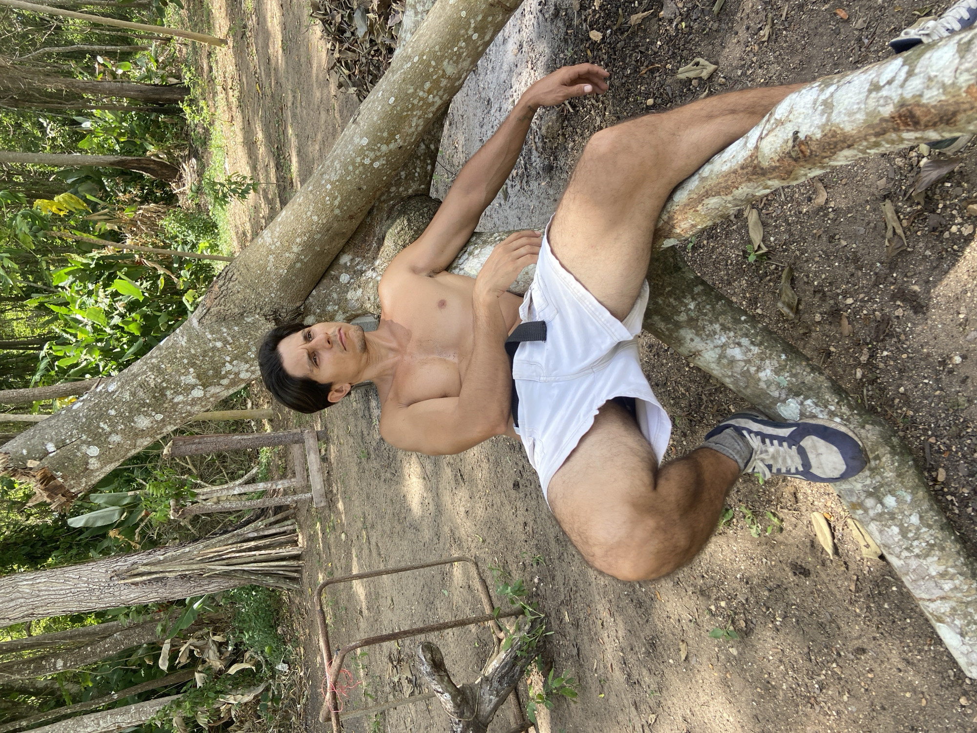 In Los Velásquez, Venezuela, a shirtless man sits casually on a fallen tree trunk in a wooded area. He is wearing white shorts and athletic shoes. The man is looking off to the side with a pensive expression. The background consists of lush green foliage and more trees, suggesting a natural, outdoor setting. The ground is covered in dirt and fallen leaves. There is a rusty metal structure to the left of the man.